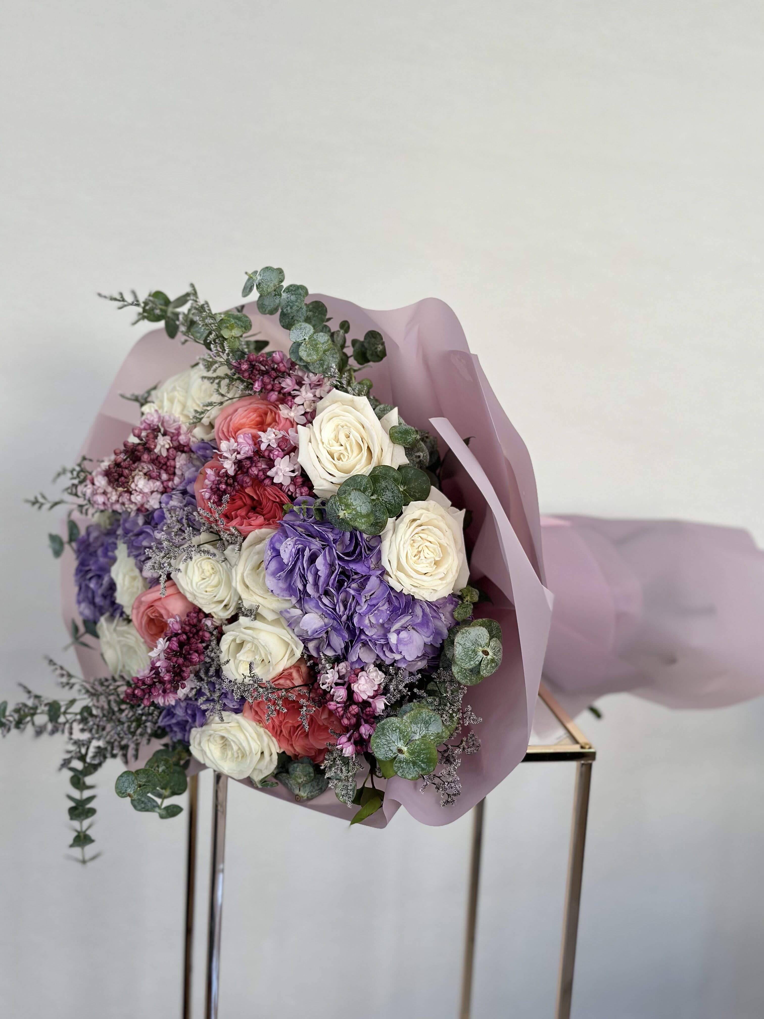 Lilac Garden Bouquet - Indulge in the elegance of this bouquet, where pristine white roses, vibrant coral garden roses, rich purple hydrangeas, and delicate lilac blooms come together in perfect harmony. Each flower whispers tales of sincerity and warmth, creating a captivating ensemble that celebrates beauty and grace. Note: Lilacs are seasonal and will be substituted when out of season.   