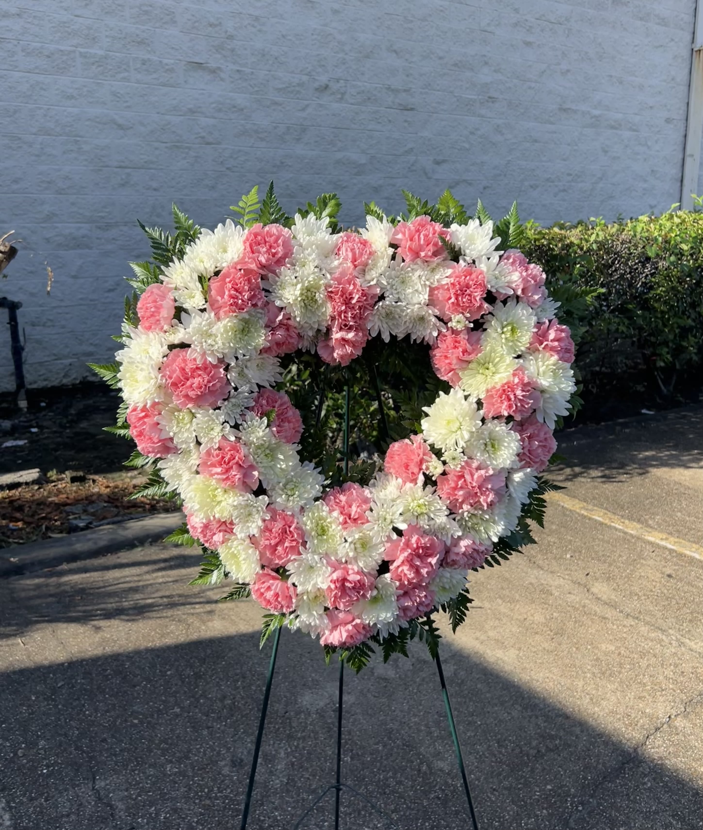Pink Heart/Corazon Rosa - This elegant heart-shaped standing wreath features a soft blend of pink and white carnations, accented with lush greenery. Designed to express heartfelt sympathy and everlasting love, it serves as a beautiful tribute to honor a cherished memory.  Approximate 18'' Open Heart Wreath  Esta delicada corona en forma de corazón combina claveles rosados y blancos, realzada con follaje fresco. Es un arreglo lleno de ternura y amor eterno, ideal para rendir homenaje a la memoria de un ser querido.  Aproximadamente 18&quot; Corona abierta en forma de corazon 