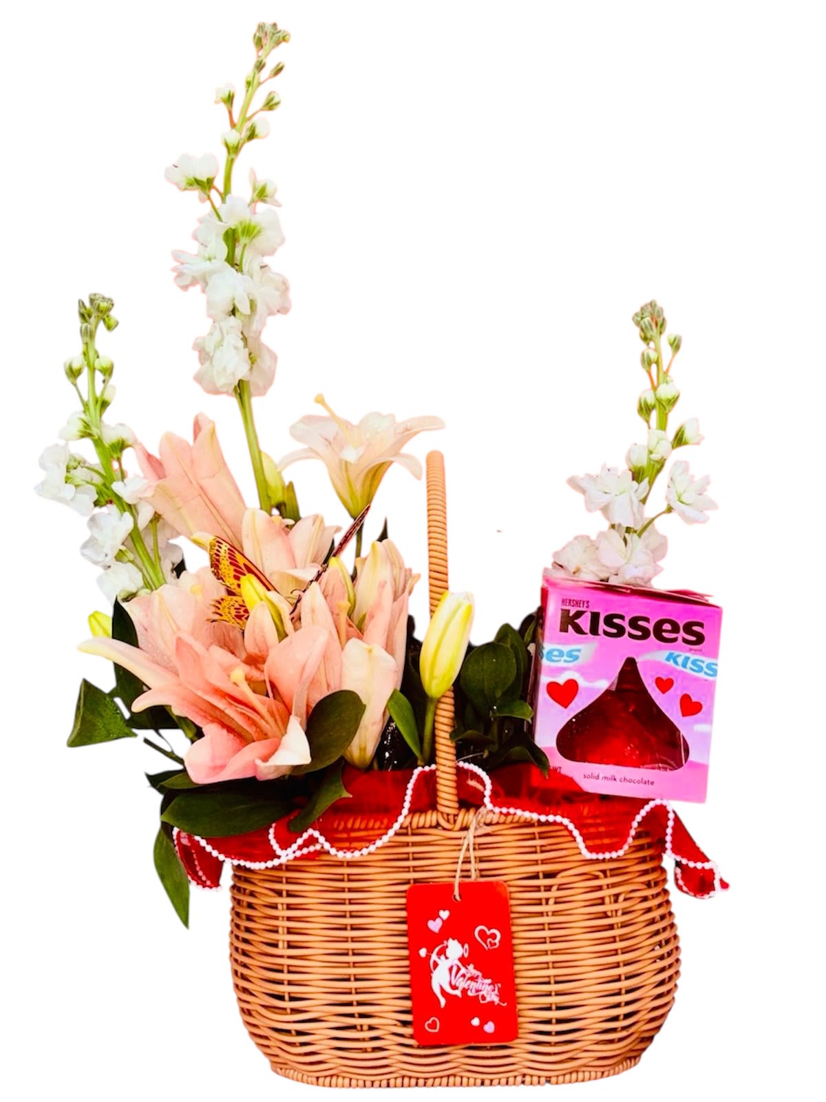 Valentine's Cradle - This charming gift basket is a cozy embrace of sweetness and affection. Nestled inside a wicker cradle lined with red fabric and laced trim are delicate pink and white lilies, complemented by other soft white blooms. A box of Hershey's Kisses adds a deliciously romantic touch, echoing the theme of love and indulgence. The red Valentine’s Day tag with heart motifs seals it as a delightful celebration of heartfelt connection—perfect for gifting someone you cherish.
