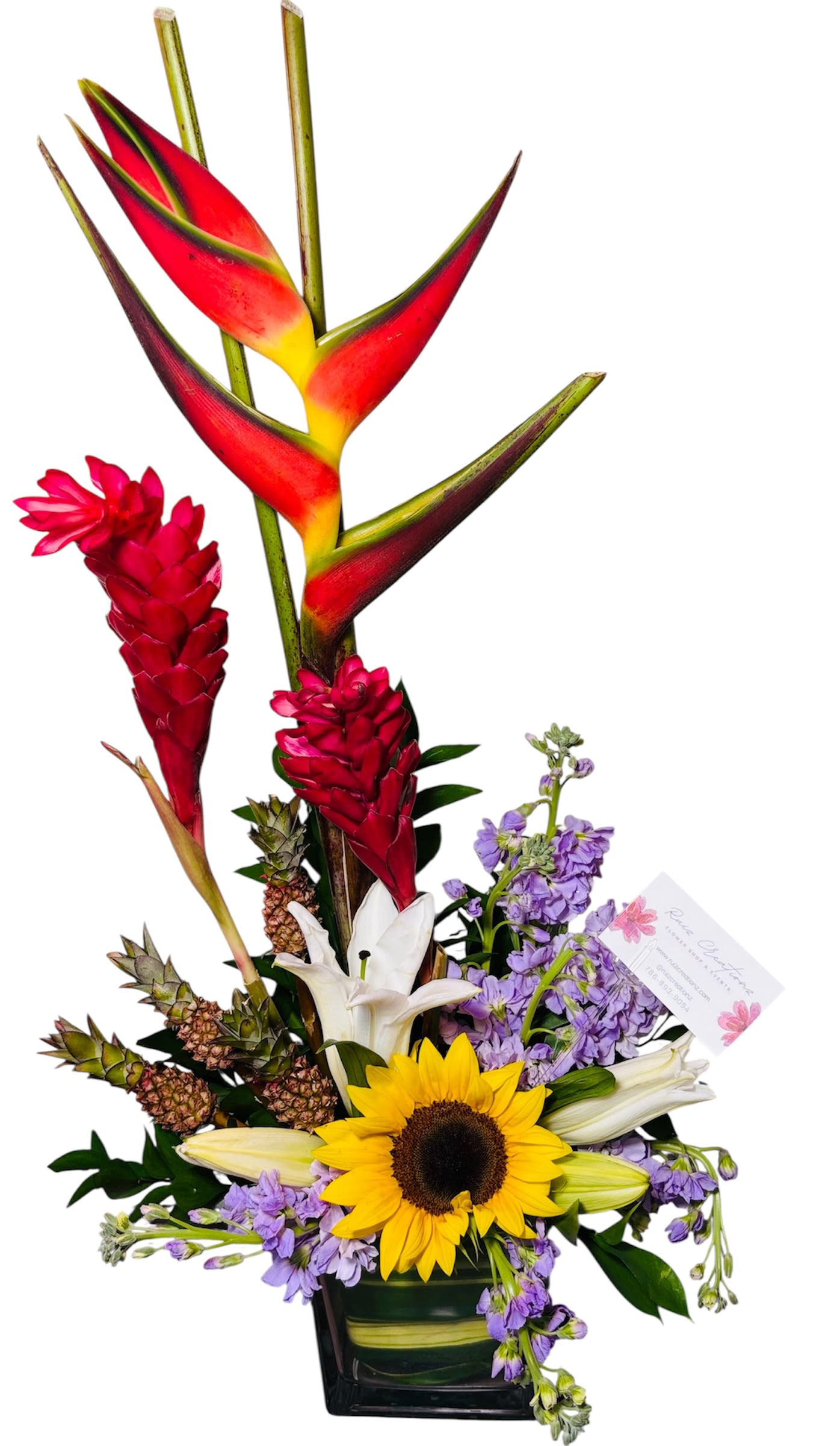 Tropical Muse - This dynamic arrangement is an artistic burst of tropical boldness and joyful gratitude. Vibrant heliconia and red ginger flowers rise dramatically, framed by creamy white lilies and a sunflower that radiates golden warmth. Pineapple plants lend playful texture, while purple accents soften the composition with a whisper of elegance. Tucked neatly inside a clear glass vase, the arrangement is paired with a branded card from “Rose Chapter,” adding a stylish, personalized touch.  It’s a striking fusion of tropical vibrance and heartfelt appreciation—ideal for events, thank-you gestures, or brand-forward displays that celebrate nature’s diversity.