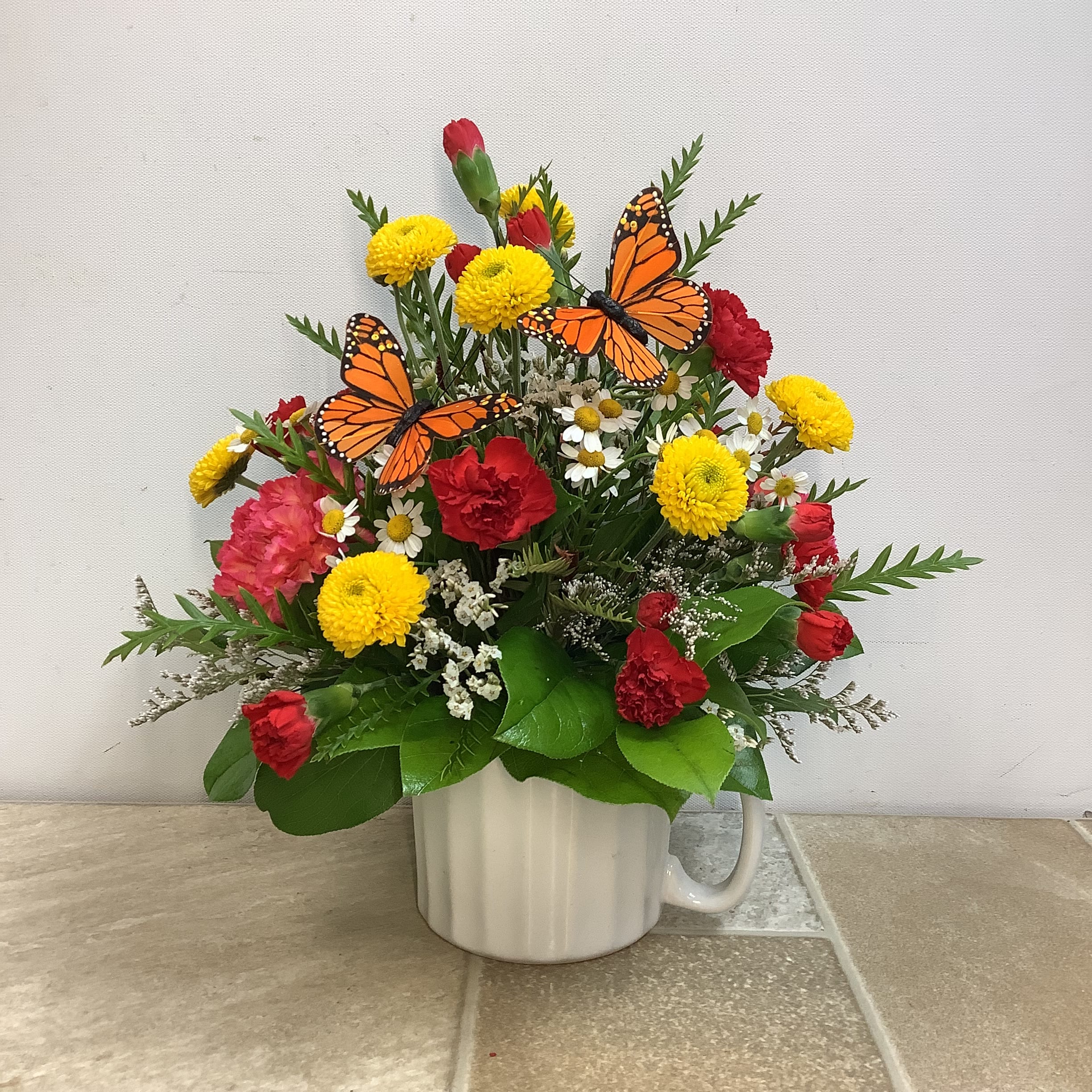 Monarch Medley - Yellow button mums, pink carnations, chamomile and red mini carnations beautifully combine in this charming fresh arrangement. Delivered in a reusable white ceramic mug, our Monarch Medley bouquet is sure to brighten your recipient’s day. It features 2 monarch butterflies and measures 12” x 12”.