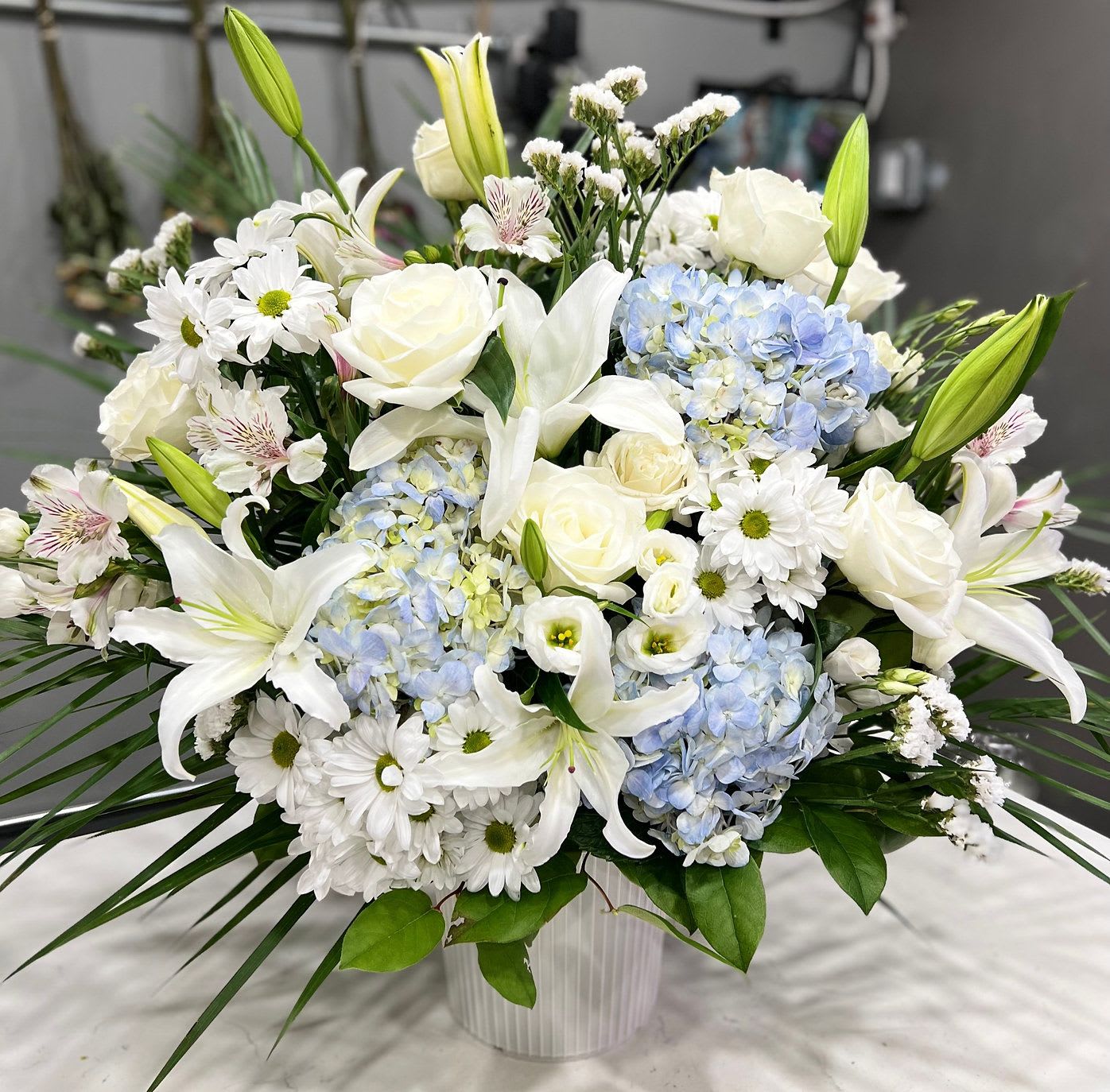 Heartfelt Blue Floor Basket - Graceful white lilies, soft blue hydrangeas, and creamy roses bloom together in this serene arrangement, echoing the sky’s quiet grace. “Heartfelt Blue Floor Basket” is a peaceful farewell—perfect for memorials, sympathy offerings, or gentle moments of reflection.  For when words fall short, and only beauty remains.