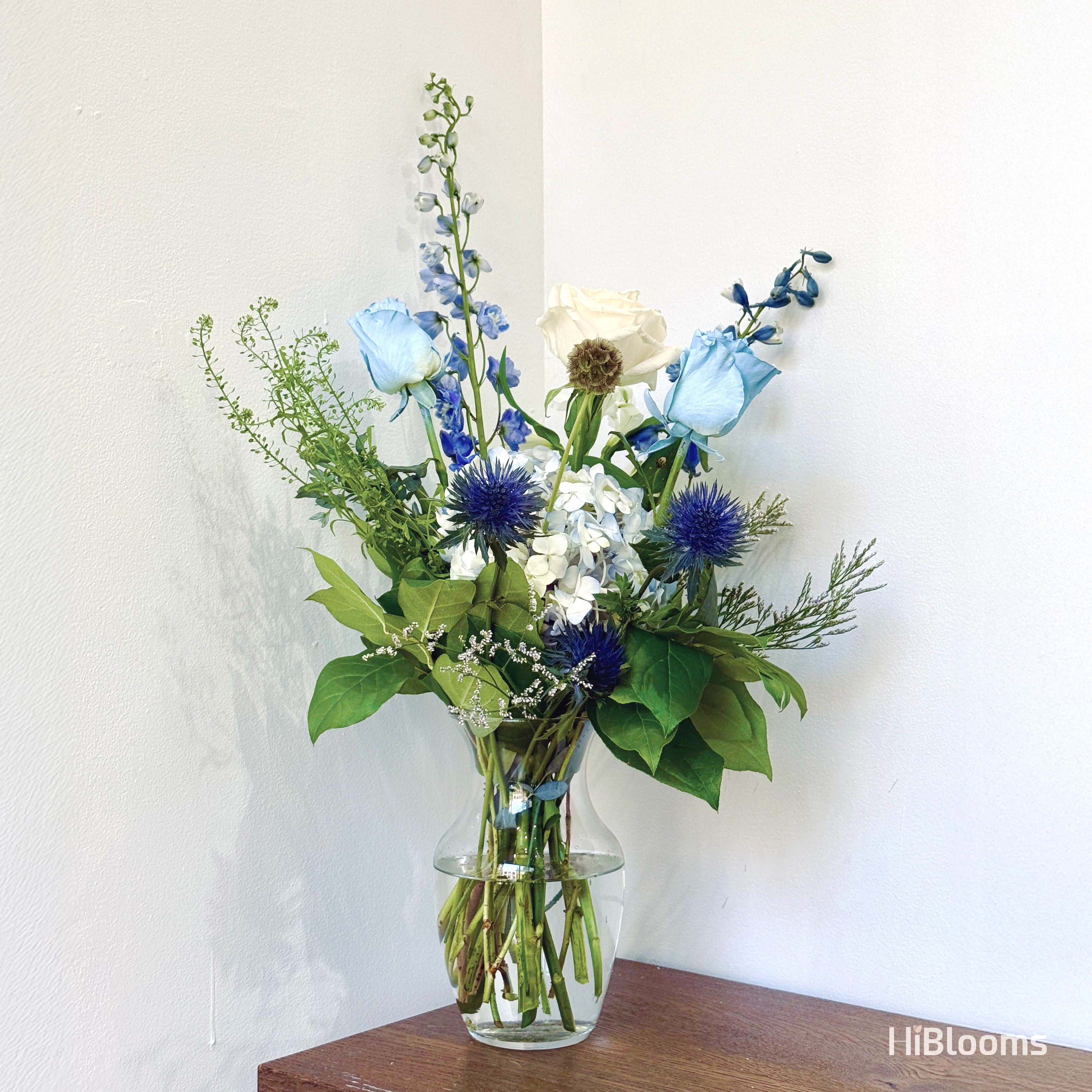 Lilylight Reverie - A quiet homage to Monet’s Water Lilies, this bouquet captures the hush of light on still water. Delicate blue delphiniums and soft-dyed roses mirror the fleeting hues of dusk reflecting across a lily pond, while snowy hydrangeas evoke floating blooms. Accents of thistle and cedar lend a painterly depth, like shadows beneath the surface.  Designed for those who find poetry in reflection, Lilylight Reverie is floristry as a brushstroke—fleeting, elegant, and steeped in tranquility.   Cool-toned, impressionist-inspired  Vase not included | Approx. 16-18&quot; tall  Part of the HiBlooms x Art History Collection