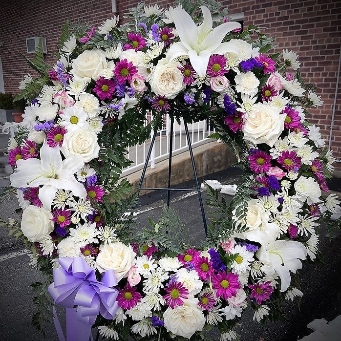 Purple Funeral Wreath - This standing wreath blends pure white blooms with rich violet and lavender tones—symbolizing peace, dignity, and deep remembrance. “Purple Funeral Wreath” speaks to the strength of love that endures, and the grace of a life well remembered. Finished with a soft ribbon and structured greens, it’s a touching expression of goodbye.  For those who were both gentle and strong, luminous and lasting.