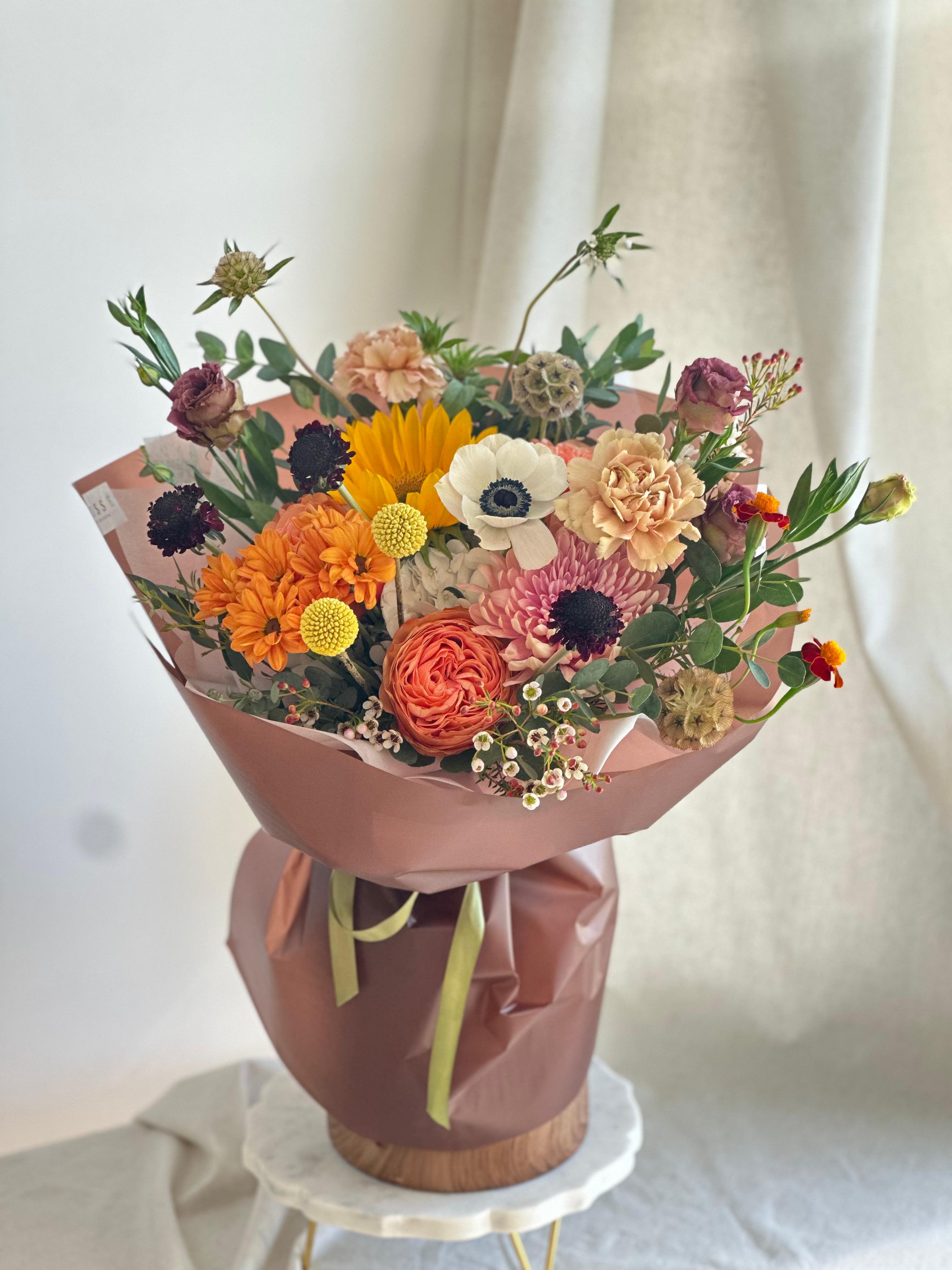 Fall in Love  - A round bouquet with yellow, orange, green, dark tones, and white flowers, wrapped in brown paper.   Flower types may vary depending on the season.
