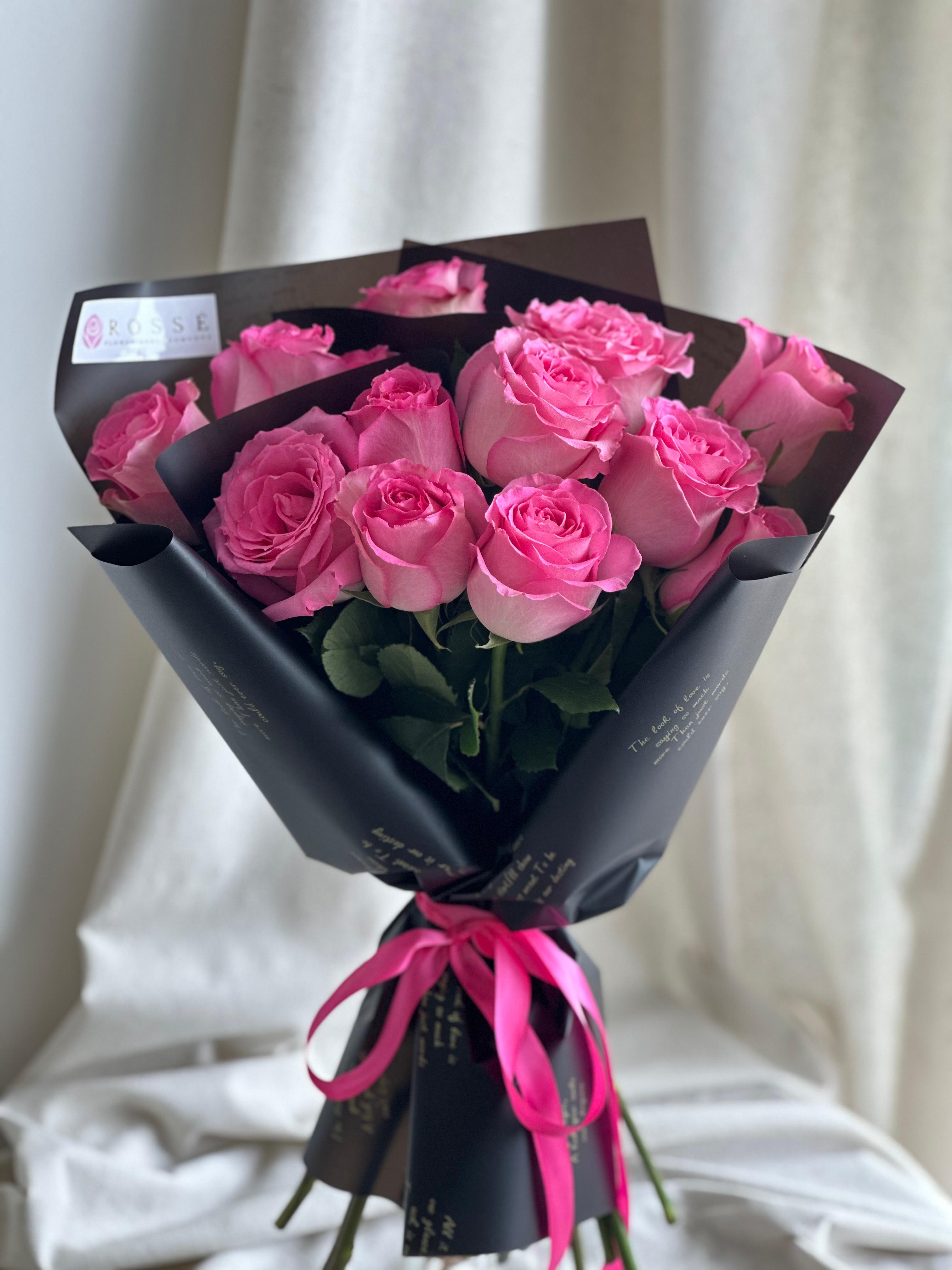 Rosey in Pink  - A front-facing bouquet of roses with layered paper accents, wrapped in black and gold paper.   Flower types may vary depending on the season.