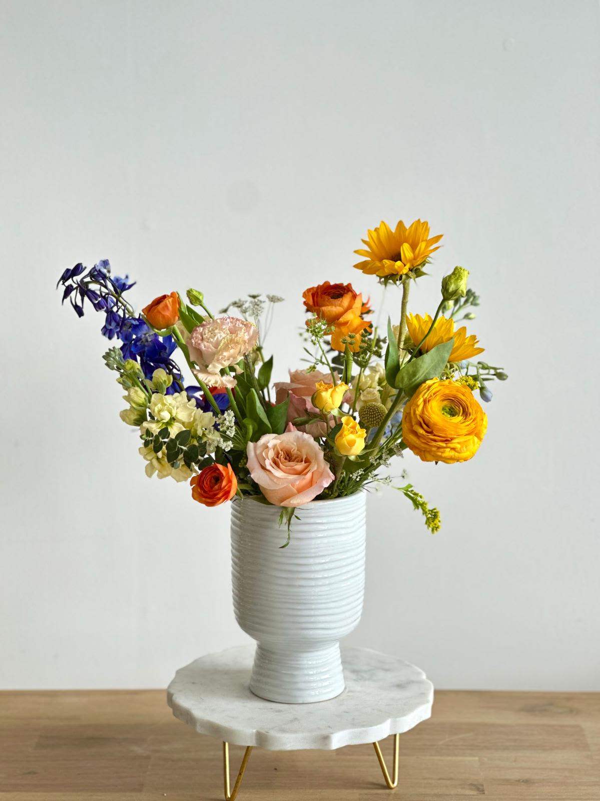 Vibrant Summer - A medium white vase with a front-facing arrangement of blue, orange, and yellow flowers.   Flower types may vary depending on the season.