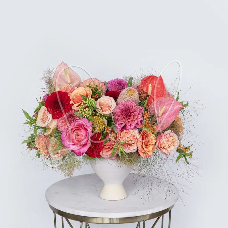 Good Vibrations - A stunning arrangement which turns up the good vibrations of summer in pinks, corals + reds  Featuring: Dahlias, Anthuriums, Garden Roses &amp; Protea in a white ceramic vase. 