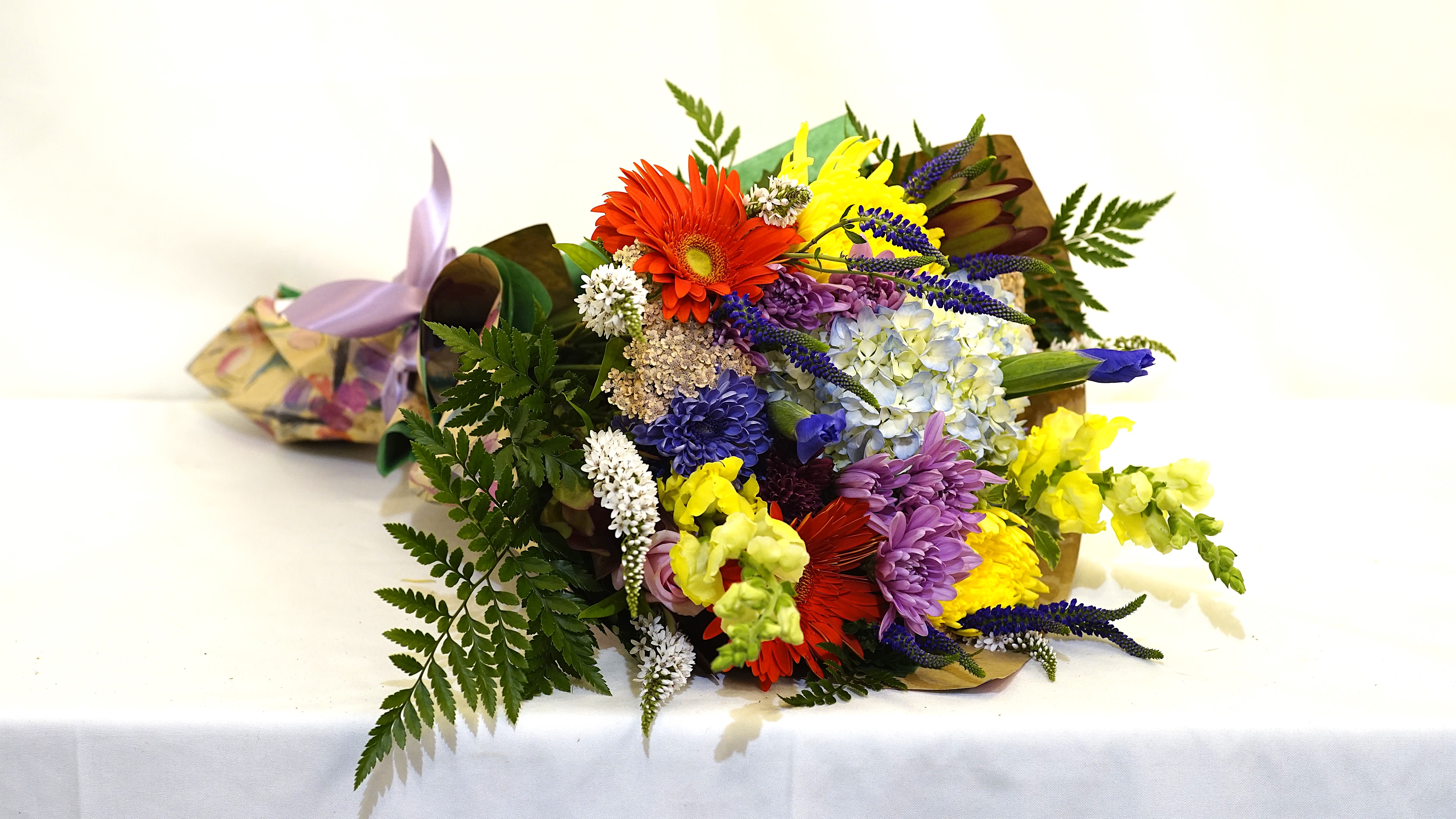 Radiant Joy - A bright burst of happiness in every bloom, Ideal for gifting or as a vibrant tabletop display. The Radiant Joy Hand-Tied Bouquet is a lively, garden-style mix designed to celebrate life’s vibrant moments. With a playful balance of color, texture, and fragrance, this bouquet is perfect for birthdays, thank-you gifts, celebrations, or simply to brighten someone’s day.  Hand-tied bouquet, wrapped in kraft and floral paper with a satin ribbon finish. Size: Approx. 14&quot;–16&quot; diameter Color Palette: Bright tones of yellow, orange, lavender, purple, white, and green.  *SYMPATHY/FUNERAL: PLEASE NOTE THE TIME, PLACE, &amp; NAME OF THE DECEASED FOR THE SERVICE IN THE SPECIAL NOTES SECTION OF YOUR ORDER - WE ARE UNABLE TO GUARANTEE DELIVERY TIME WITHOUT THIS.*