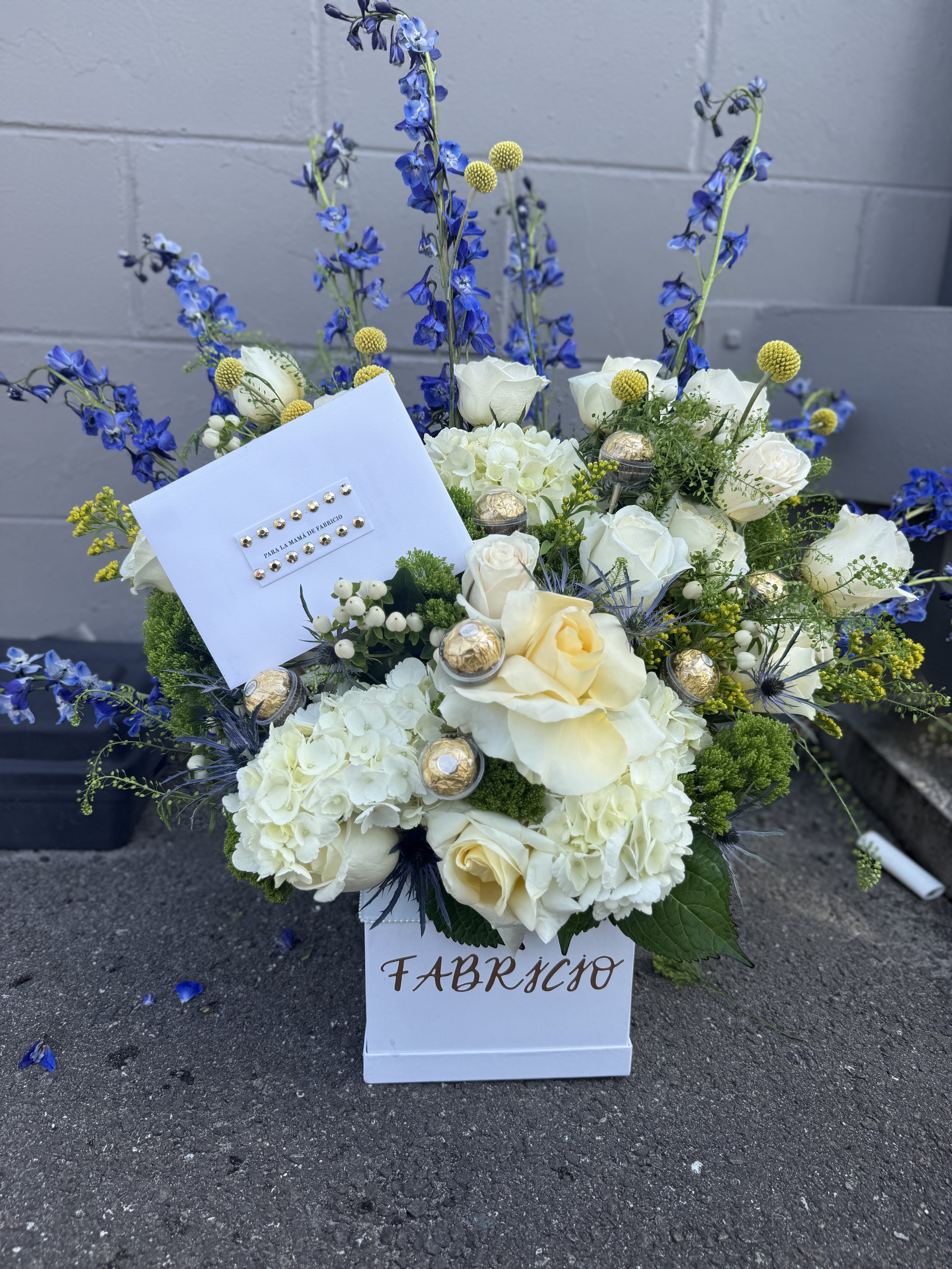 XXL Golden Serenity XXL Golden Serenity - An elegant and sophisticated arrangement designed to celebrate someone truly special. Featuring a harmonious blend of ivory and soft yellow roses, lush white hydrangeas, and textured accents of greenery and blue thistle, this piece radiates calm and grace. Delicate Craspedia balls and tall violet delphiniums add a modern and striking contrast, while golden Ferrero Rocher chocolates offer a sweet touch of luxury. Presented in a clean white box, this arrangement is perfect for birthdays, congratulations, or simply to honor someone with refined taste.