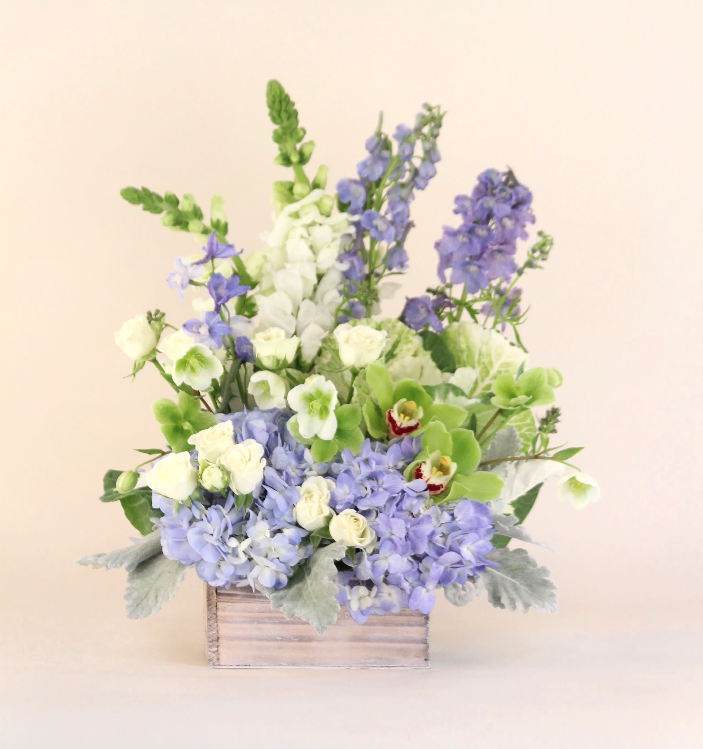 Blue Sky - A lush arrangement of blues and whites in a white wash wooden box.  Flowers may include hydrangeas, spray roses, delphinium, hellebore, snapdragon and orchids.  Wooden box dimension: (7&quot; W x 4&quot; H)