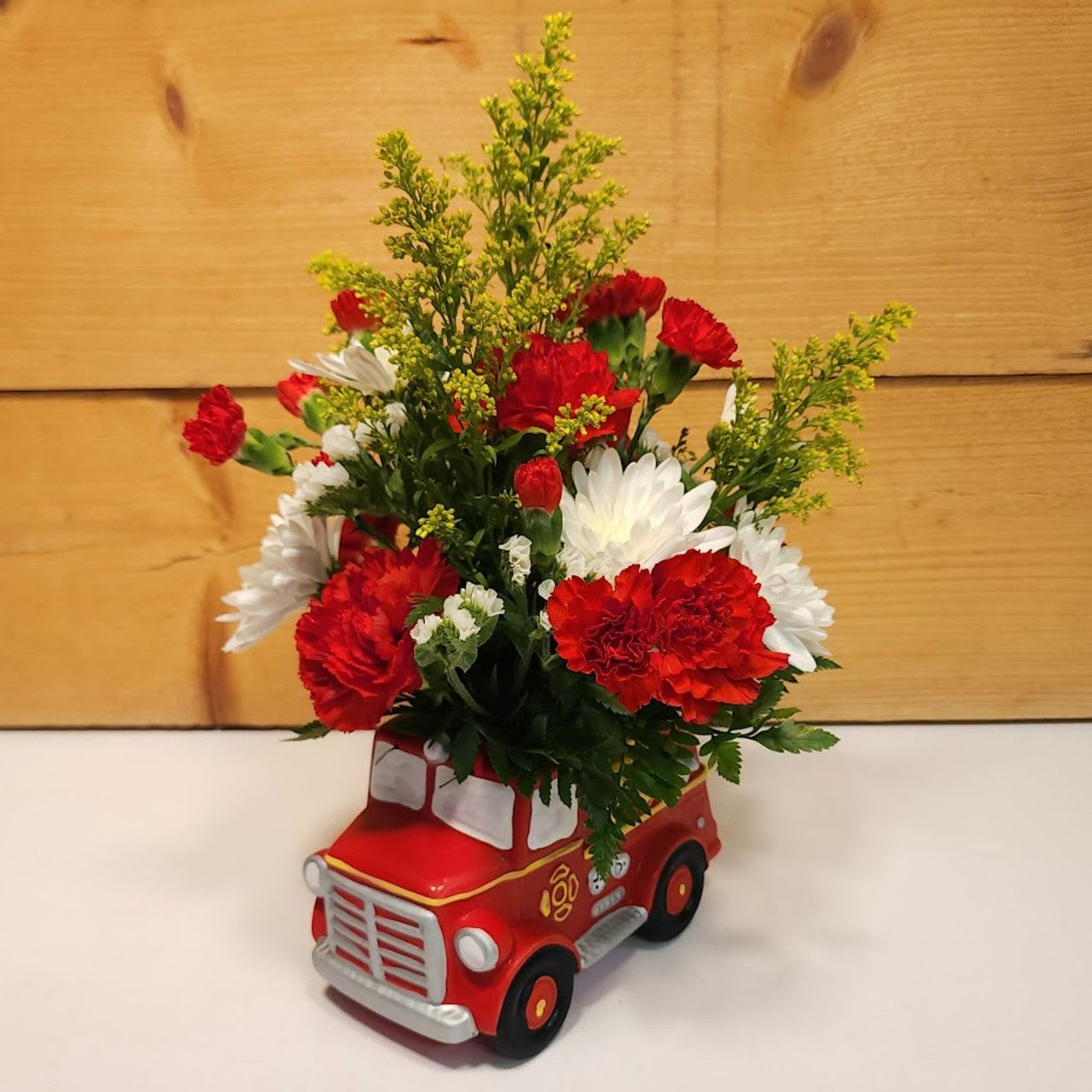 Firetruck Florals Fiesta - Celebrate the arrival of a little one with this charming floral arrangement presented in a vibrant red ceramic firetruck container. Brimming with fresh and joyful blooms, this design is perfect for welcoming a new baby with a burst of color and cheer.  The arrangement features pristine white cremones that symbolize purity and new beginnings, bright yellow solidago adding a touch of sunny optimism, and vivid red carnations that bring a sense of excitement and joy. The firetruck container adds a playful and whimsical touch, making this a delightful centerpiece for any baby shower or new baby celebration.  Standing as a symbol of joy and fun, this arrangement combines the classic charm of a firetruck with the beauty of fresh flowers, creating a heartwarming tribute to the newest member of the family.