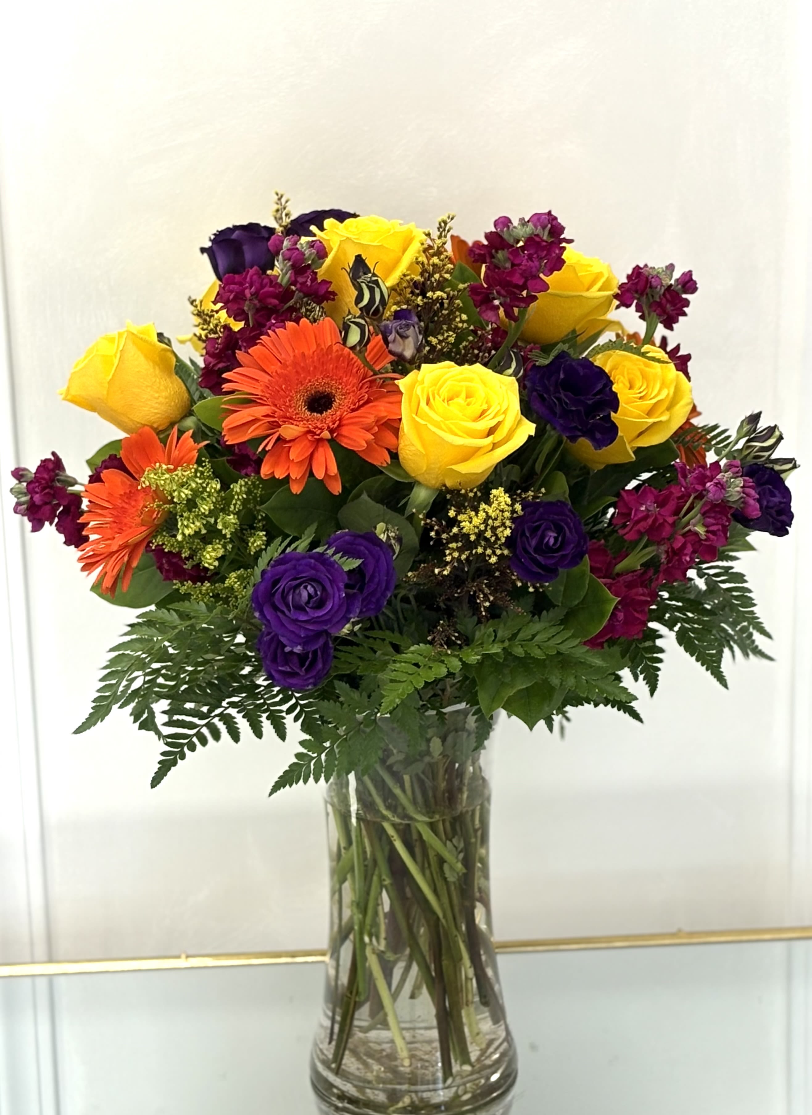 Festival of Joy By VLM Flowers - A true celebration in bloom, this bouquet bursts with festive energy through radiant yellow roses, bright orange gerberas, rich purple lisianthus, and magenta stock flowers. Arranged in a clear vase with fresh greenery, it’s a perfect pick-me-up that brings color and cheer wherever it goes.  Perfect For: Birthday surprises, celebrations, or sending sunshine and smiles.