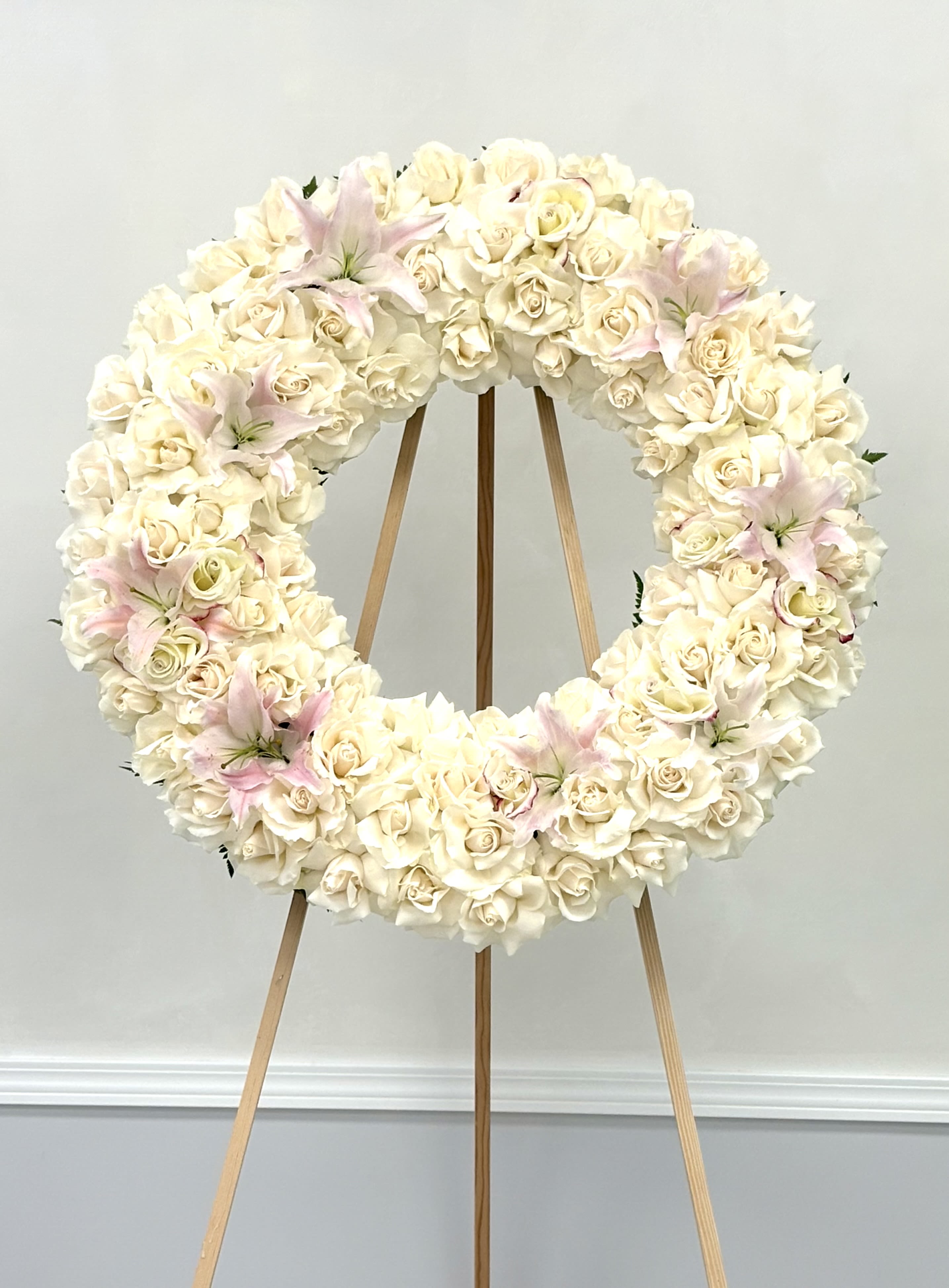 Silent Grace By VLM Flowers - This all-white wreath, delicately accented with pale pink lilies, speaks in the language of quiet elegance and enduring love. The soft textures and muted tones offer a comforting presence during a time of loss.