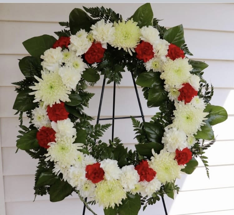 Loving Memory wreath - A circular funeral wreath is a timeless tribute, its unbroken shape symbolizing eternal love and remembrance. Imagine it gracefully adorned with creamy white roses, fragrant lilies, and delicate baby’s breath, woven together in soft harmony. Nestled between are accents of silver eucalyptus leaves or ivy, adding texture and a touch of natural elegance.  Touches of pale blue hydrangea or soft lavender blooms may be interspersed for a gentle, calming effect—subtle hues that whisper peace and serenity. The arrangement rests on a simple easel, allowing it to stand with quiet dignity, a floral embrace that honors a life beautifully lived.  It’s the kind of wreath that feels like a gentle farewell and a heartfelt thank-you wrapped into one. Would you like help with the message for the banner or card that accompanies it? We are happy to help you. 