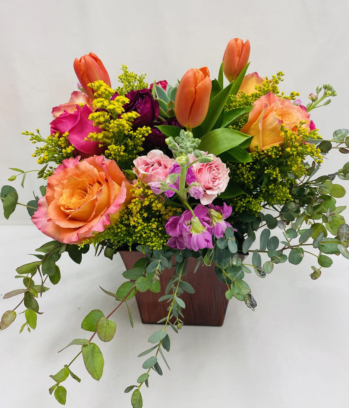 Obsessed  - Centrally placed succulent surrounded by brightly colored selection of flowers including Roses, Safari, Snap Dragon, Solidago, and eucalyptus leaves, in a square wood like vase.