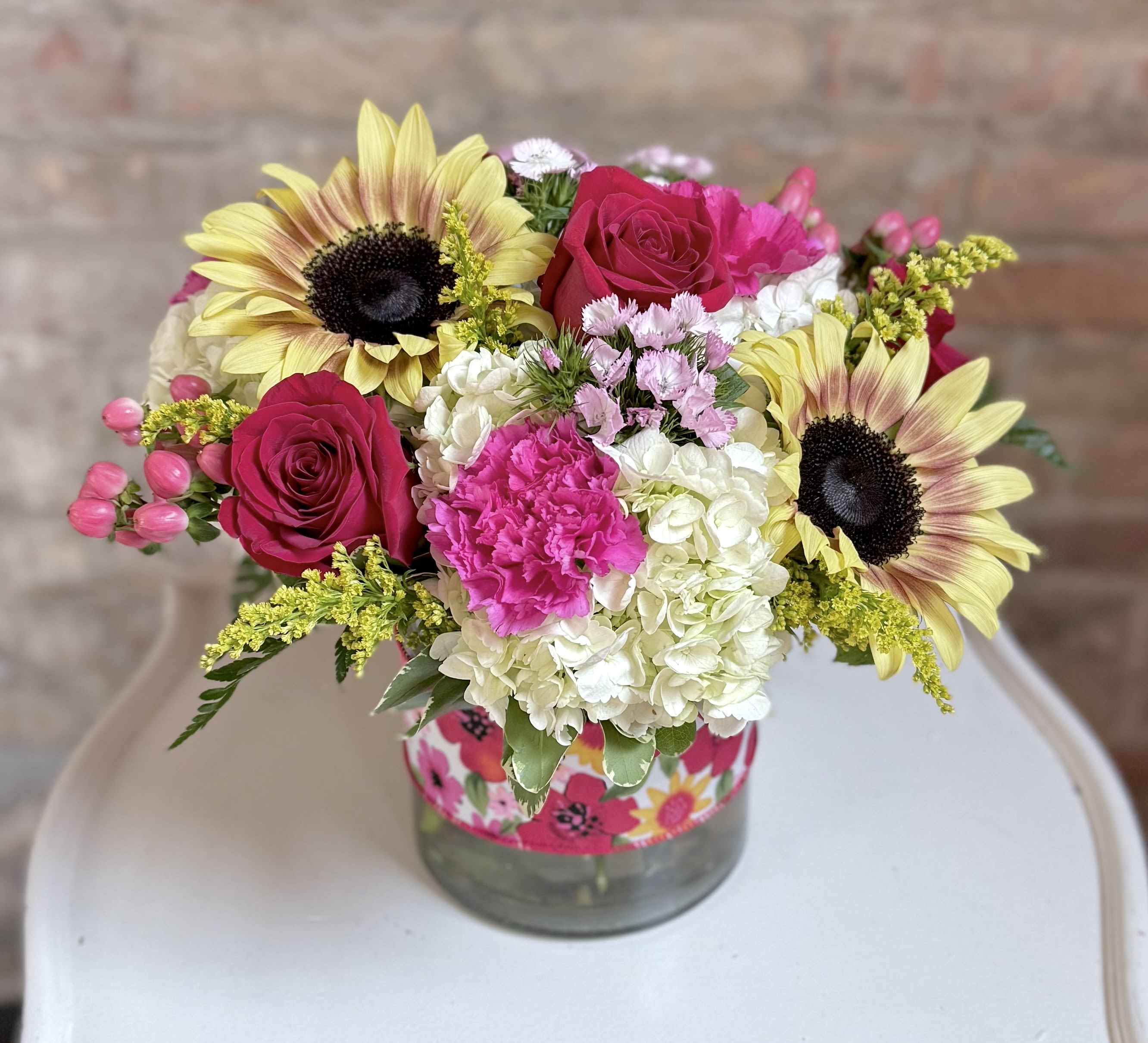Quinn Quinn - A low wide cylinder accented with a bright summer floral ribbon. The cylinder is filled with vibrant red, pink, coral, yellow and white flowers! Including roses, sunflowers, berries, hydrangeas and carnations. Very bright and perfect for summer!