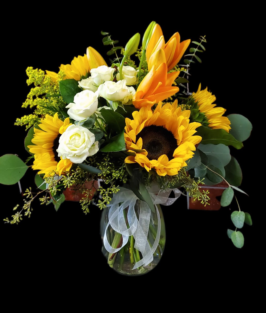 Sunflower Surprise - A one sided flower arrangement designed with fresh flowers to include sunflowers, orange lilies, white spray roses, filler, and mixed greens in a vase. With a festive yellow bow.  * Some stages of flower life may vary. (Example: Lilies may be more open than imaged.)  Flower shortages and product availability means that some substitutions will apply. Value will always be filled. This bouquet might include eucalyptus amongst the greenery. It may be toxic to pets including but not limited to Cats and Dogs if digested.