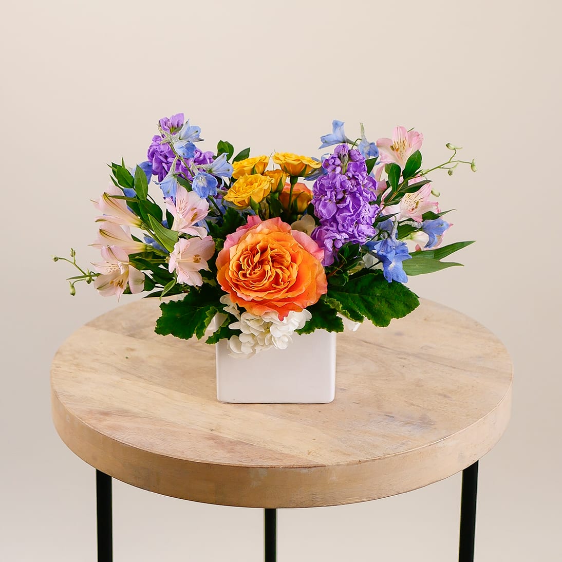 Sunset Meadow - Brighten any room with Sunset Meadow, a fresh flower arrangement bursting with vibrant color and seasonal charm. This handcrafted floral design features an eye-catching Free Spirit garden rose, yellow spray roses, pink alstroemeria, lavender stock, blue delphinium, and white hydrangea. Set in a sleek white ceramic container, it’s a modern floral arrangement perfect for birthdays, get well wishes, thank-you gifts, or home decor. Ideal for anyone who loves colorful flowers and contemporary design, Sunset Meadow adds a cheerful touch to any space. Approximately measures 10 inches tall and 12 inches wide with flowers.