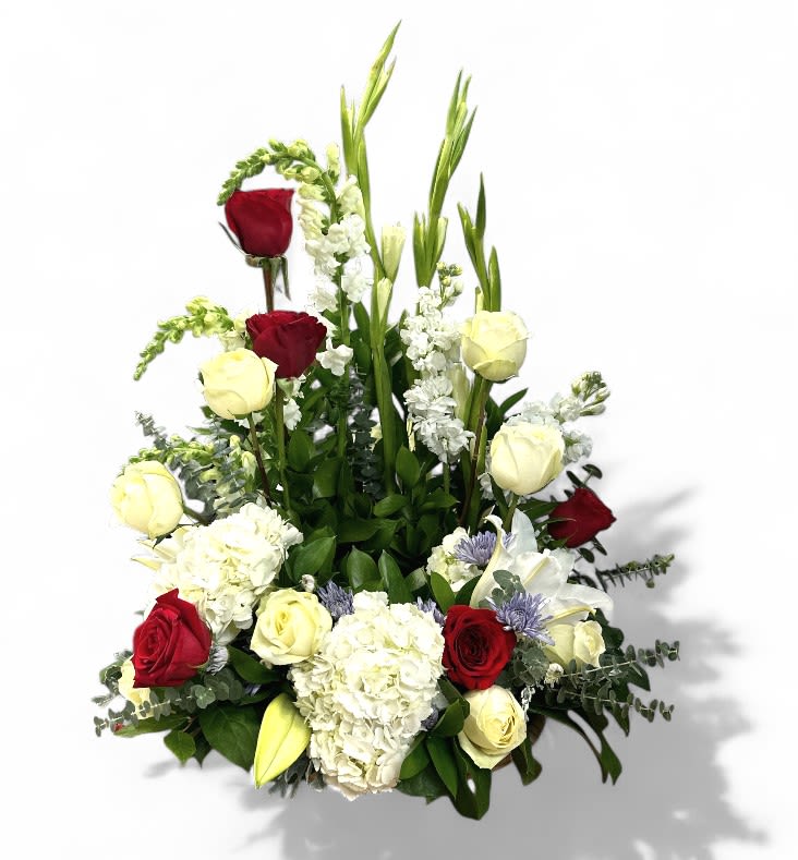 Freedom's Valor Urn Tribute - The Freedom's Valor Urn Surround is a breathtaking tribute that beautifully embodies patriotism and honor. Crafted with a harmonious blend of vibrant and elegant blooms, this surround features tall white gladiolas and delicate white snapdragons that symbolize purity and remembrance. Surrounding these are lush white hydrangeas and white stock sticks, adding fullness and softness to the arrangement. Accentuating the design are striking red and white roses, representing sacrifice and unity, complemented by the regal blue mums that evoke a sense of steadfast loyalty. Elegant Asiatic lilies lend a touch of grace, while the mixed green foliage and eucalyptus provide a rich, natural backdrop, enhancing the overall patriotic theme. This stunning arrangement serves as a heartfelt tribute to honor and valor, celebrating the spirit of freedom with dignity and grace.  (Urn or Photo not included.)