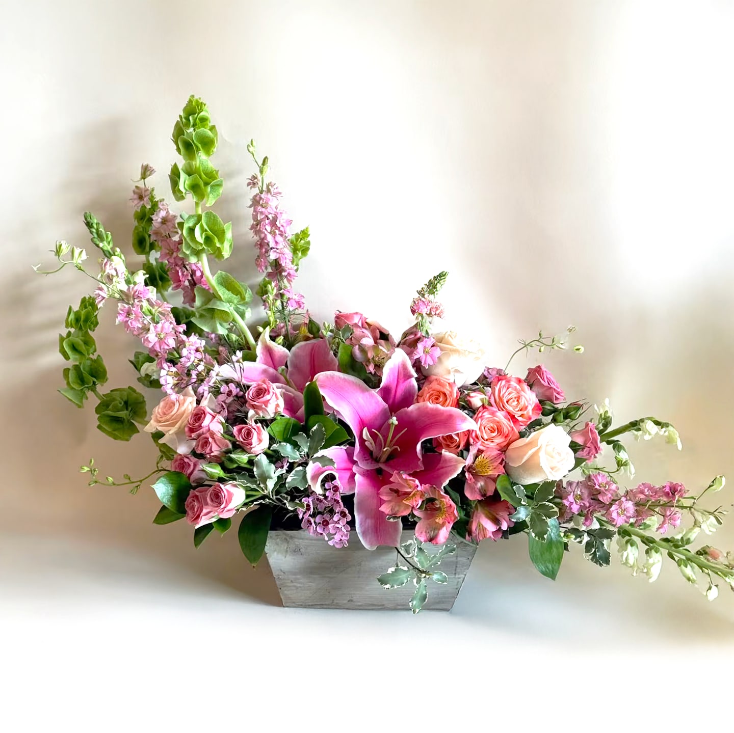 Lily Garden - Experience the delightful charm of our enchanting flower arrangement, expertly designed to showcase the natural beauty of pink lilies, delicate pink roses, bells of Ireland, and lush greenery. All arranged with care in a rustic container, this arrangement exudes an effortless grace that is sure to enchant and captivate. 