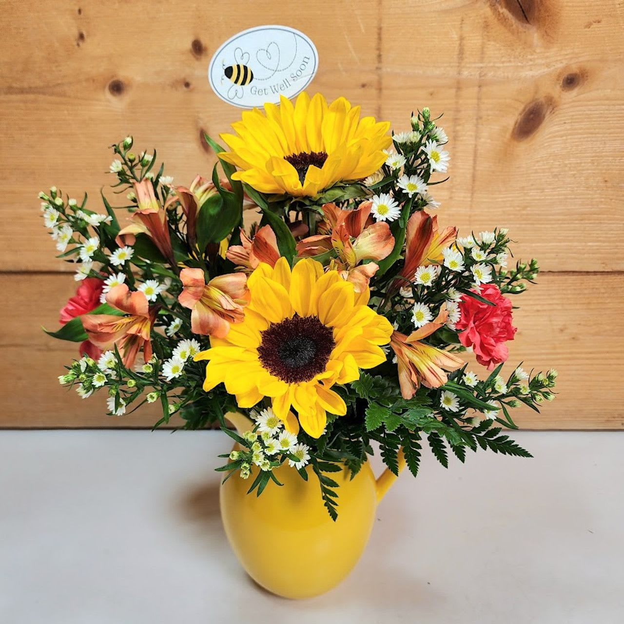 Bee Well Bouquet - The &quot;Bee Well Bouquet&quot; is a cheerful and vibrant floral arrangement designed to brighten anyone's day. This delightful bouquet features four radiant sunflowers, their golden petals symbolizing warmth and happiness. Three orange alstroemeria and three orange carnations add layers of rich, bold color, creating a joyful, lively contrast. The pristine white monte blooms offer a touch of elegance, while a whimsical &quot;get well soon&quot; bumble bee pick adds a charming, playful accent. All of this beauty is artfully arranged in a sunny yellow ceramic pitcher, standing approximately 18&quot; high and 10&quot; wide. Perfect for sending well wishes, this bouquet is a sweet and uplifting gift to bring comfort and a smile.  PLEASE NOTE: Some flowers, vases and other products pictured are subject to availability, and may be substituted for a similar item if unavailable according to our substitution policy.