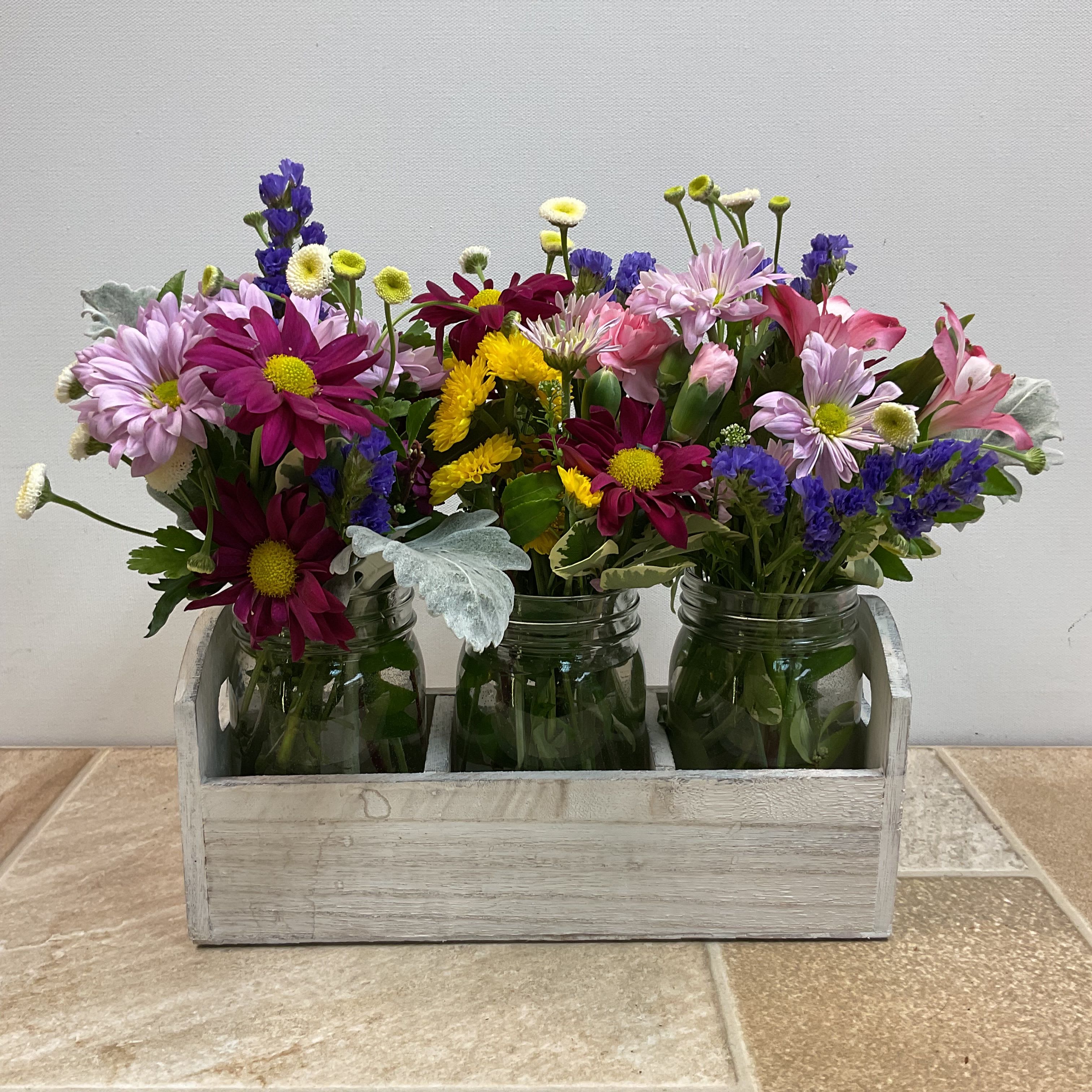 Fresh Picked Fresh Picked - This floral arrangement, called “Fresh Picked,” captures the charm of a summer meadow bouquet freshly gathered by hand. It features a lively mix of blooms -- magenta and lavender daisies, yellow button mums, pink alstroemeria, purple statice, and sweet feverfew daisies with lush greenery -- in a trio of mason jars set in a rustic whitewashed wooden caddy, giving it a relaxed, country feel. This casual and cottage-inspired design is perfect for a farmhouse table, garden party centerpiece, or cheerful gift. Approximate size: 10” wide x 9” tall. Primary flowers: daisy, mum, lily