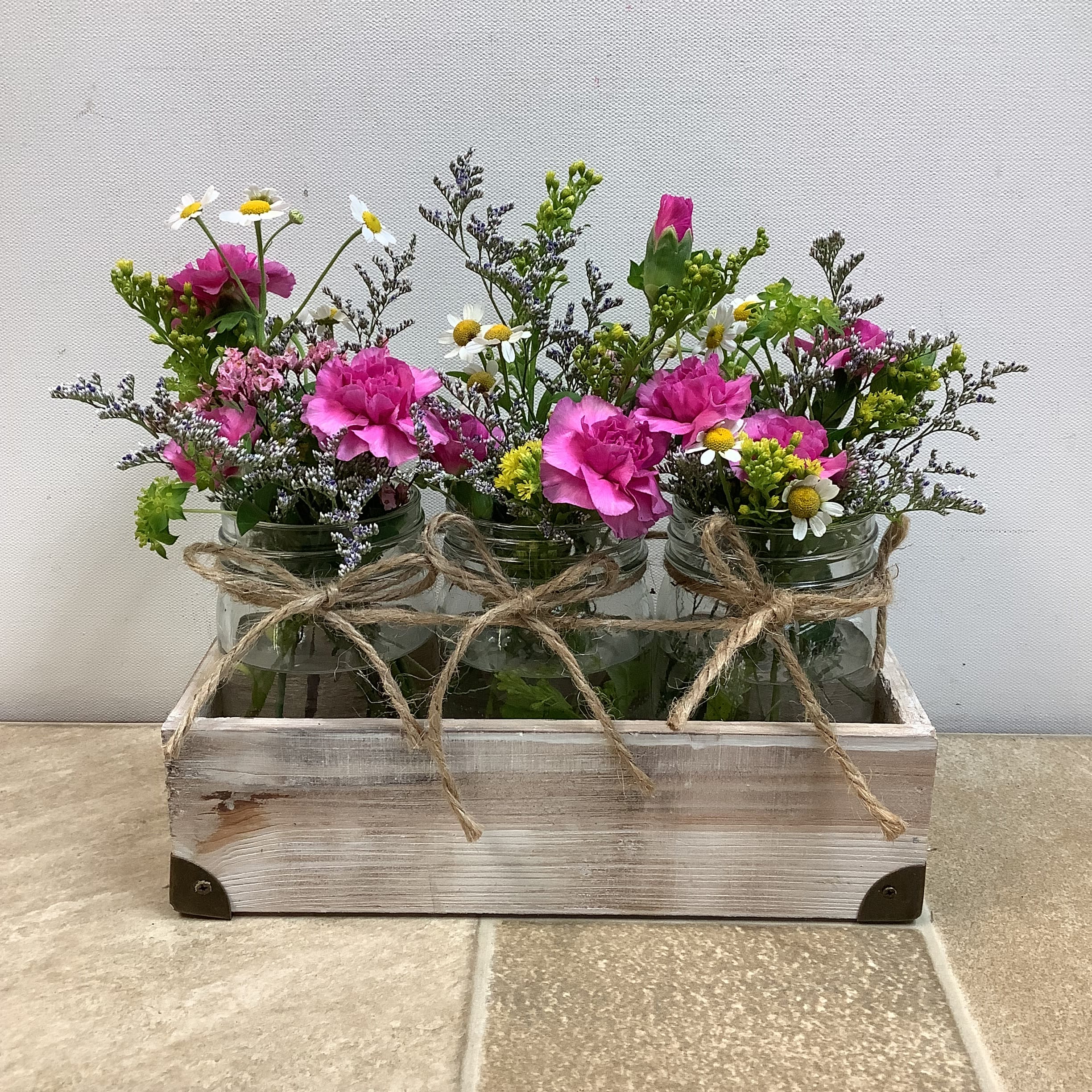 Garden Trio Garden Trio - This uniquely rustic and charming floral arrangement is presented in a white, distressed wooden box. Three clear glass mason jars, each tied with a twine bow, hold pink mini carnations, yellow solidago, and sweet daisy-like feverfew, along with pink and blue/purple limon accents and green foliage. The overall impression is fresh, natural, and homey, reminiscent of a garden or farmhouse aesthetic. Approximate size: 10” long x 5” wide x 9” tall. Primary flowers: carnation