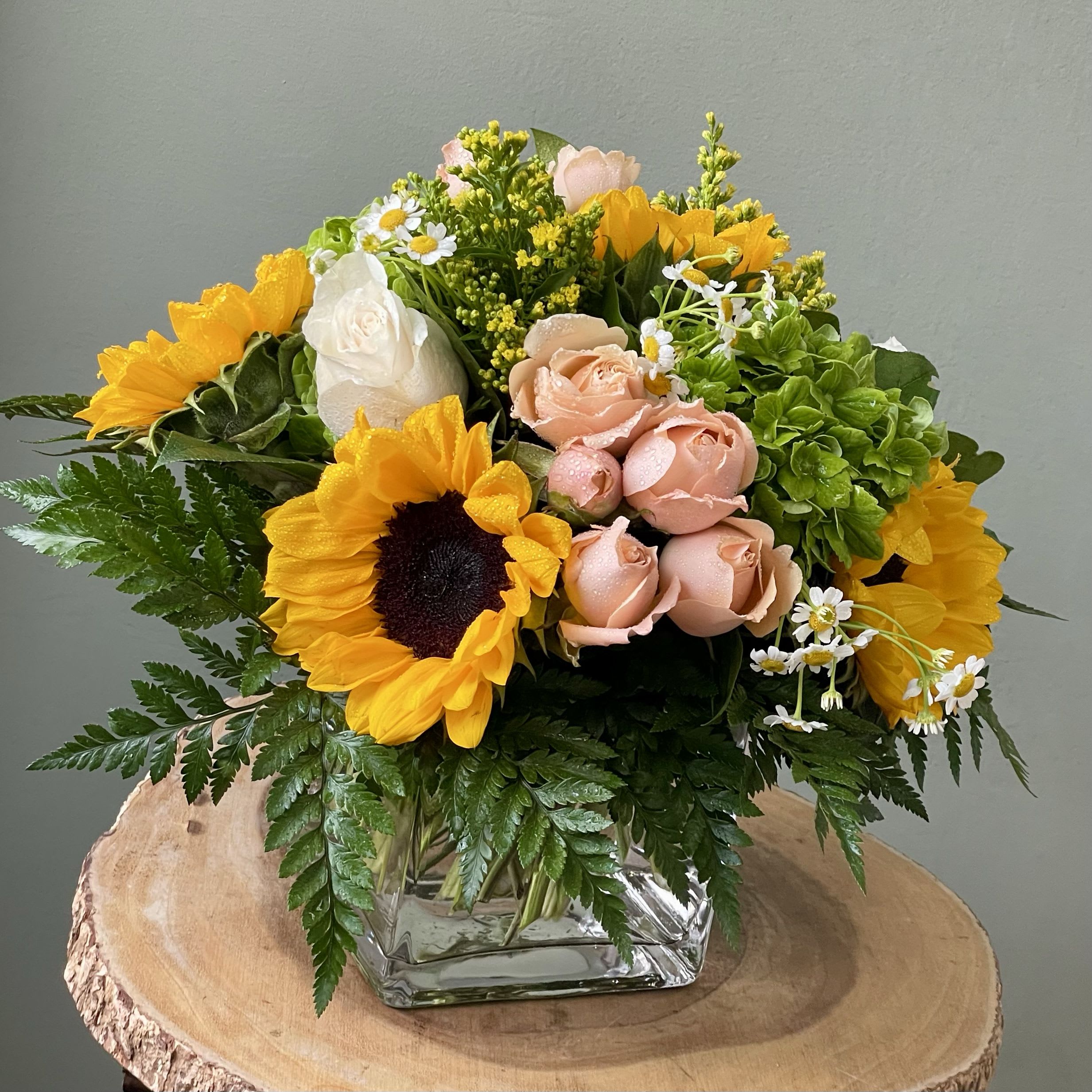 Golden Blush (SF153) - Brighten their day with this joyful, garden-fresh arrangement featuring golden sunflowers, peach spray roses, creamy white roses, green hydrangeas, and playful filler blooms like chamomile and solidago.  Designed in a modern glass cube and accented with lush greenery, this bouquet radiates warmth and cheer with every petal. Perfect for birthdays, celebrations, or just because, this arrangement brings a little sunshine to any space.