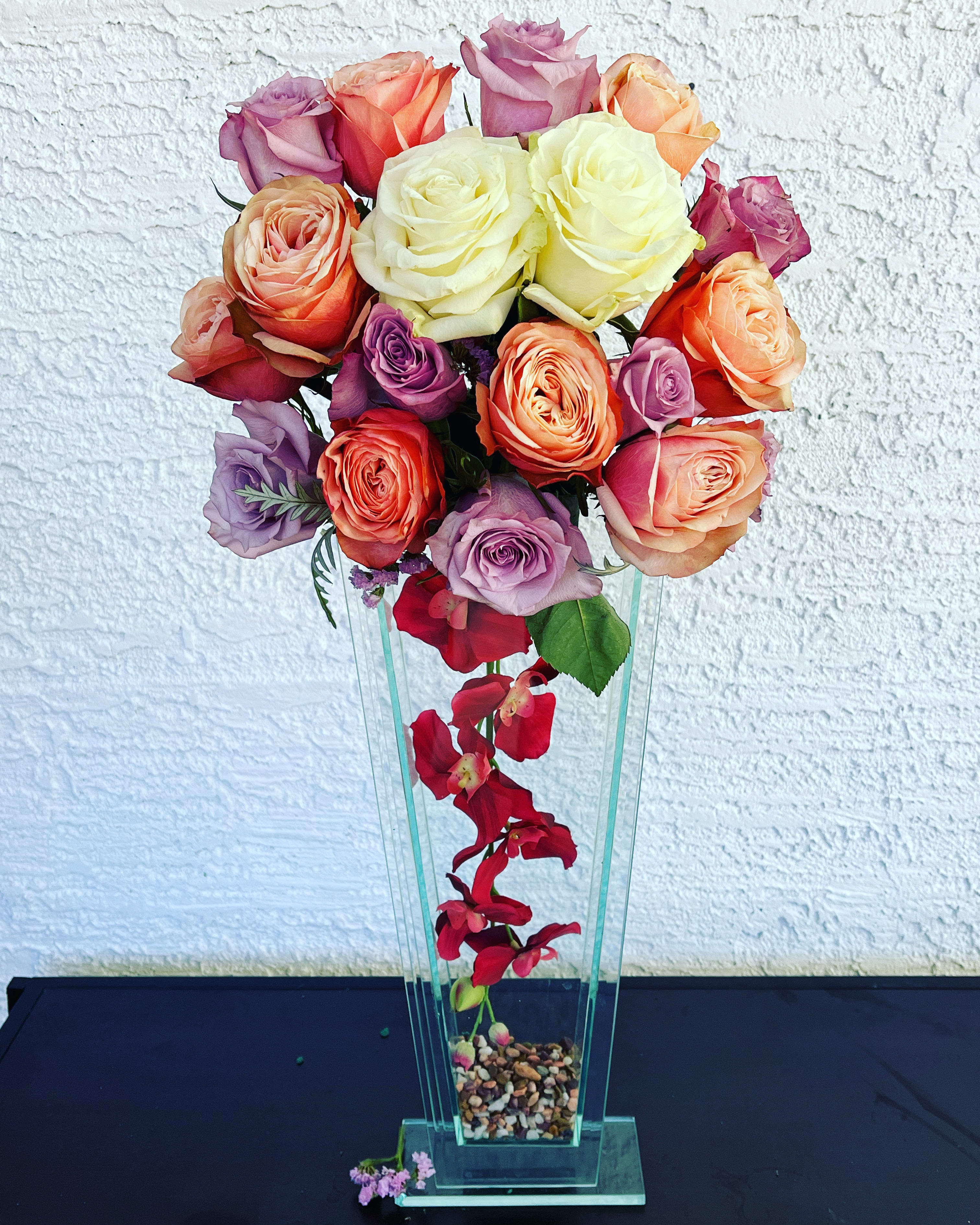 Elegant Heights - Make a stunning statement with “Elegant Heights”, a sophisticated floral display featuring a luxurious blend of lavender, peach, ivory, and coral roses arranged in a tall, tapered glass vase. A cascade of red orchids flows through the base, adding unexpected beauty and vertical elegance. Polished river stones anchor the design, giving it balance and charm.   Details: 	•	Flowers: Premium roses in pastel &amp; warm tones, red orchids 	•	Vase: Tall, modern glass tower with stone accents 	•	Style: Contemporary, romantic, upscale