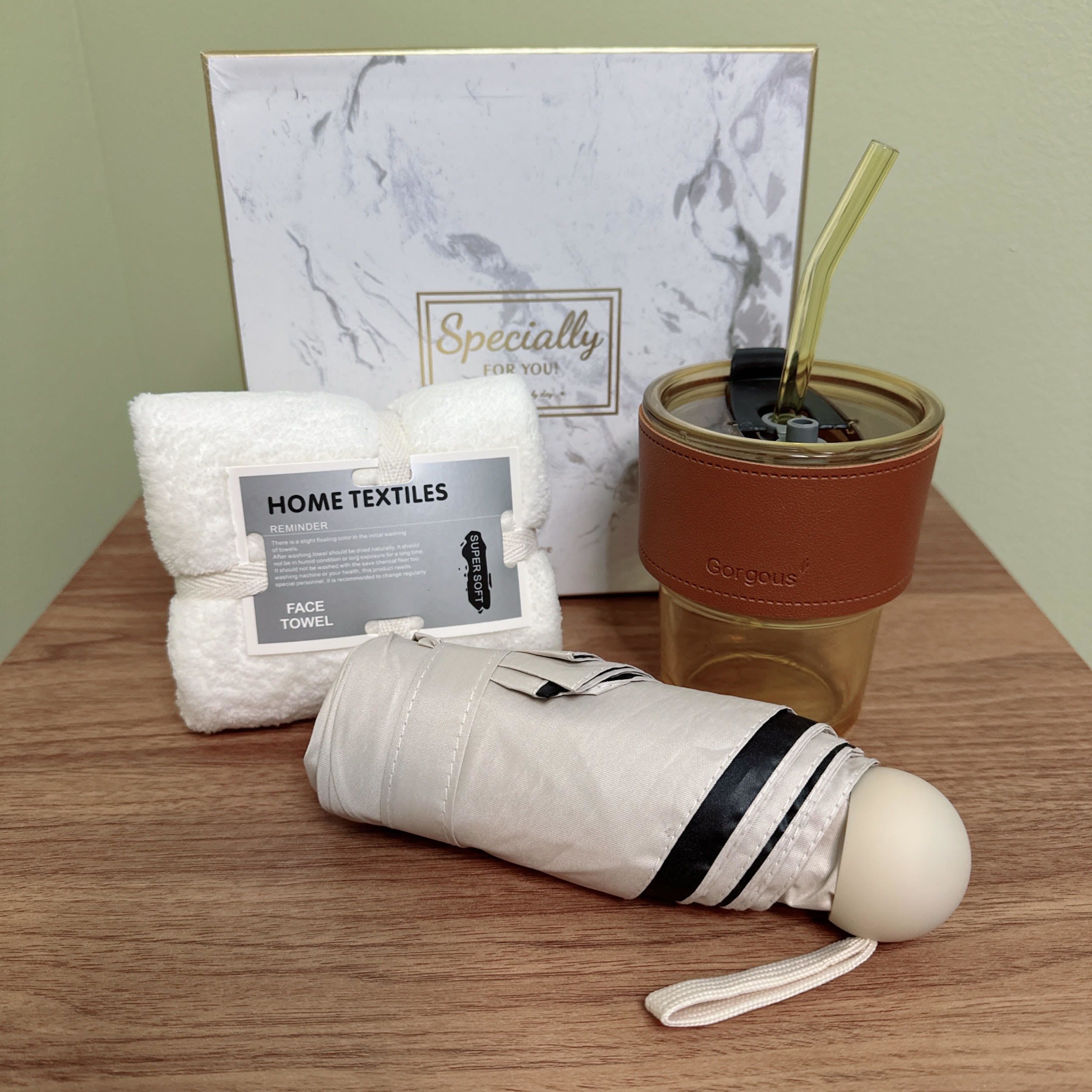 Everyday Specialty Gift Set  - This gift set is a thoughtful and practical collection of items for everyday use, presented in an attractive and elegant way.   Gift Set Includes:  1. A white face towel 2. A white umbrella with black stripes and a white handle. 3. A gold-colored tumbler with a brown leather wrap around it, featuring the word &quot;Gorgeous&quot; embossed on the front. The tumbler has a gold straw and lid.   