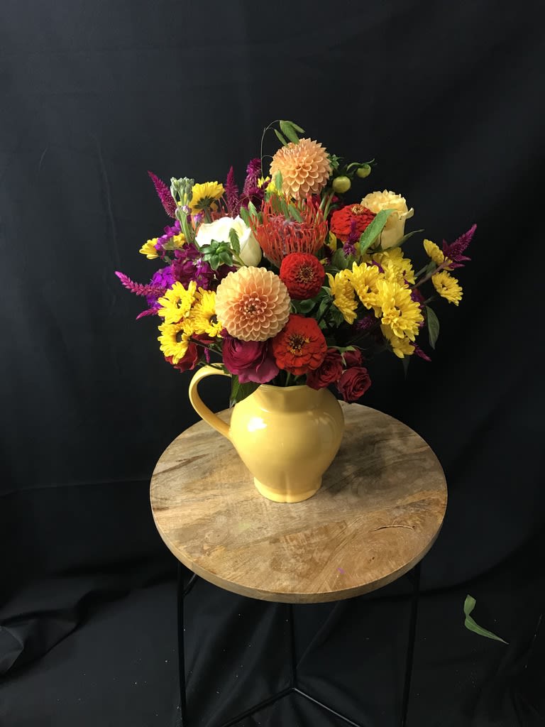Pour a Cup of Joy - Arranged in an adorable, brightly colored handle vase are some of the most vibrant blooms the season has to offer, dotted with spurts of greenery that allow for the eye to flow smoothly between every stunning blossom. 