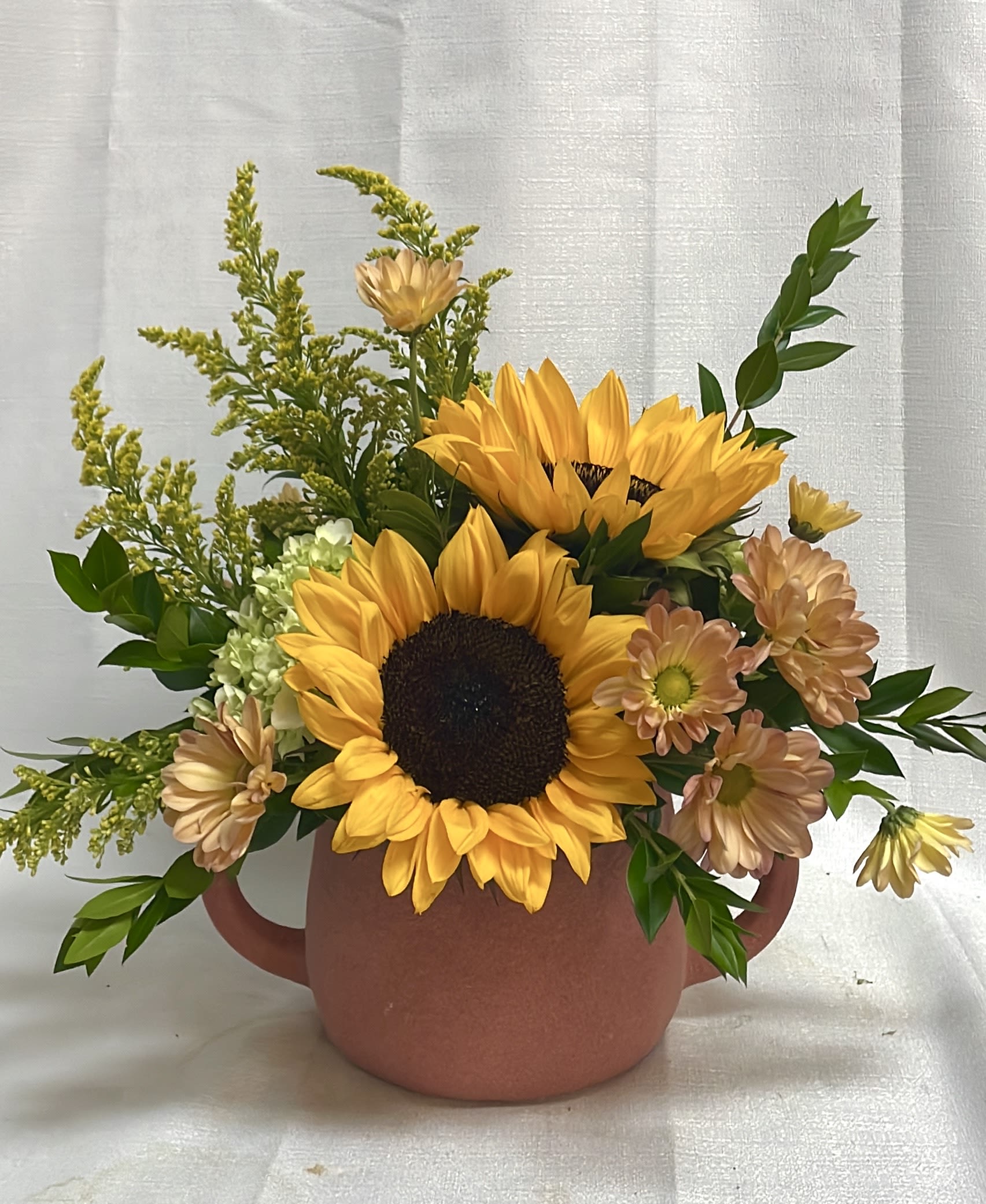 Shades of Sandstone - This gorgeous arrangement features sunflowers, orange daisies, mini green hydrangeas, and solidago in a sandy ceramic pot. Perfect for any occasion, these flowers are sure to be a beautiful centerpiece for sunny days, celebrations, or warm summer nights.