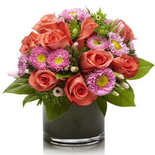 Pizazz by Sophisticated Blooms - A stunning mixed flower arrangement using vibrant coral roses and bright pink aster accented with modern green filler blooms in a chic glass vase.  • Coral Roses • Pink Matsumoto Aster • Modern Green Filler Flowers and Foliage • Glass Cube or Cylinder Vase • Personalized Gift Message • The premier luxury floral gifting experience