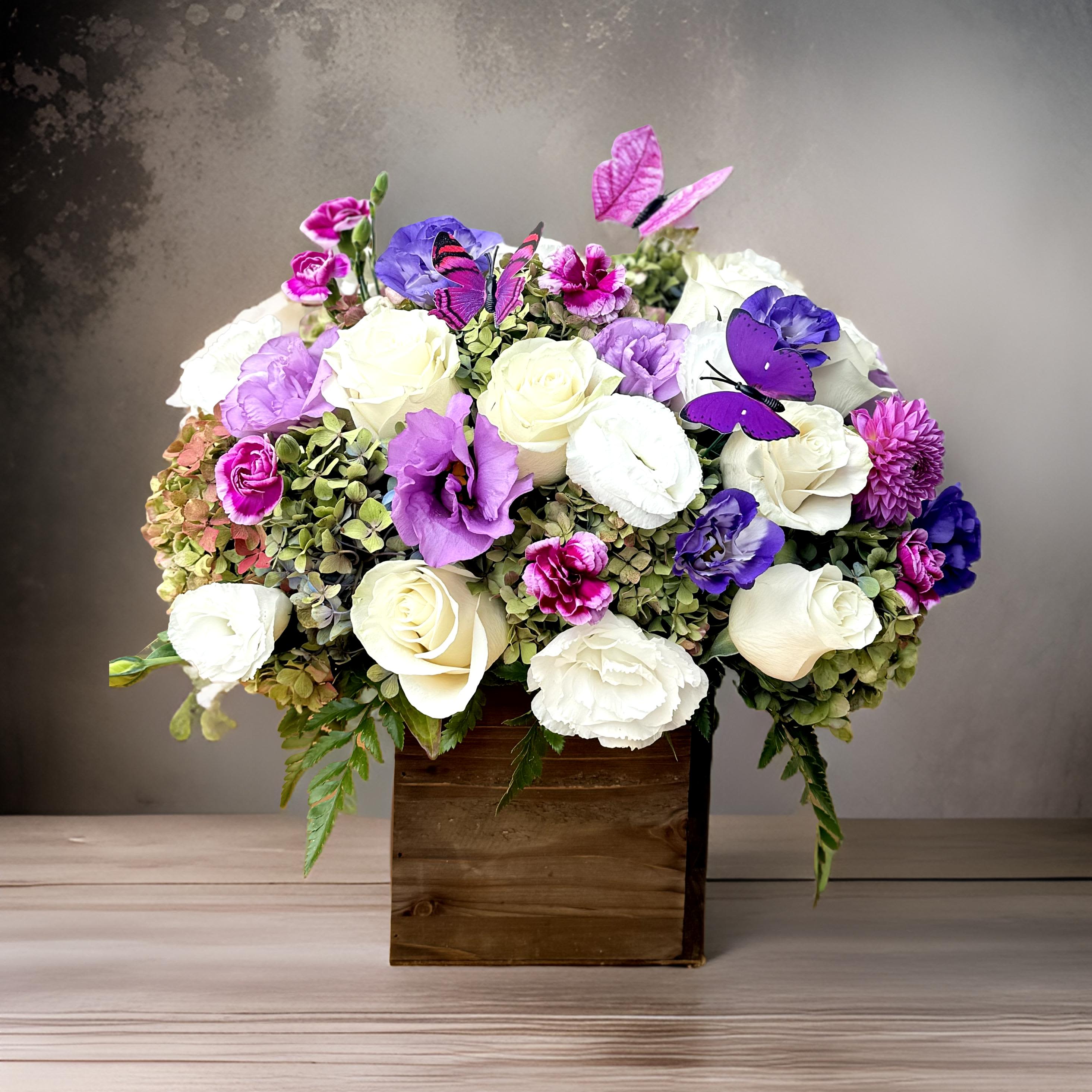 Enchanted Garden Box - This elegant floral design captures the magic of a secret garden in bloom. Nestled in a rustic wooden box, a lush composition of ivory roses, purple lisianthus, soft green hydrangeas, and violet carnations comes to life with vibrant texture and color. Finished with delicate decorative butterflies, this arrangement feels both graceful and enchanting.  The cool-toned color palette is calming yet rich, perfect for moments of celebration, appreciation, or even quiet reflection. Ideal for birthdays, sympathy gestures, housewarmings, or as a gift for someone who adores refined beauty with a whimsical twist, this piece blends natural textures with a luxurious touch.  The natural wood vessel brings warmth and earthiness, while the floral mix adds elegance and movement. Whether displayed in a modern home, a cozy office, or sent as a thoughtful gift, this arrangement offers a moment of serenity and joy.  Available for same-day flower delivery in Los Angeles.