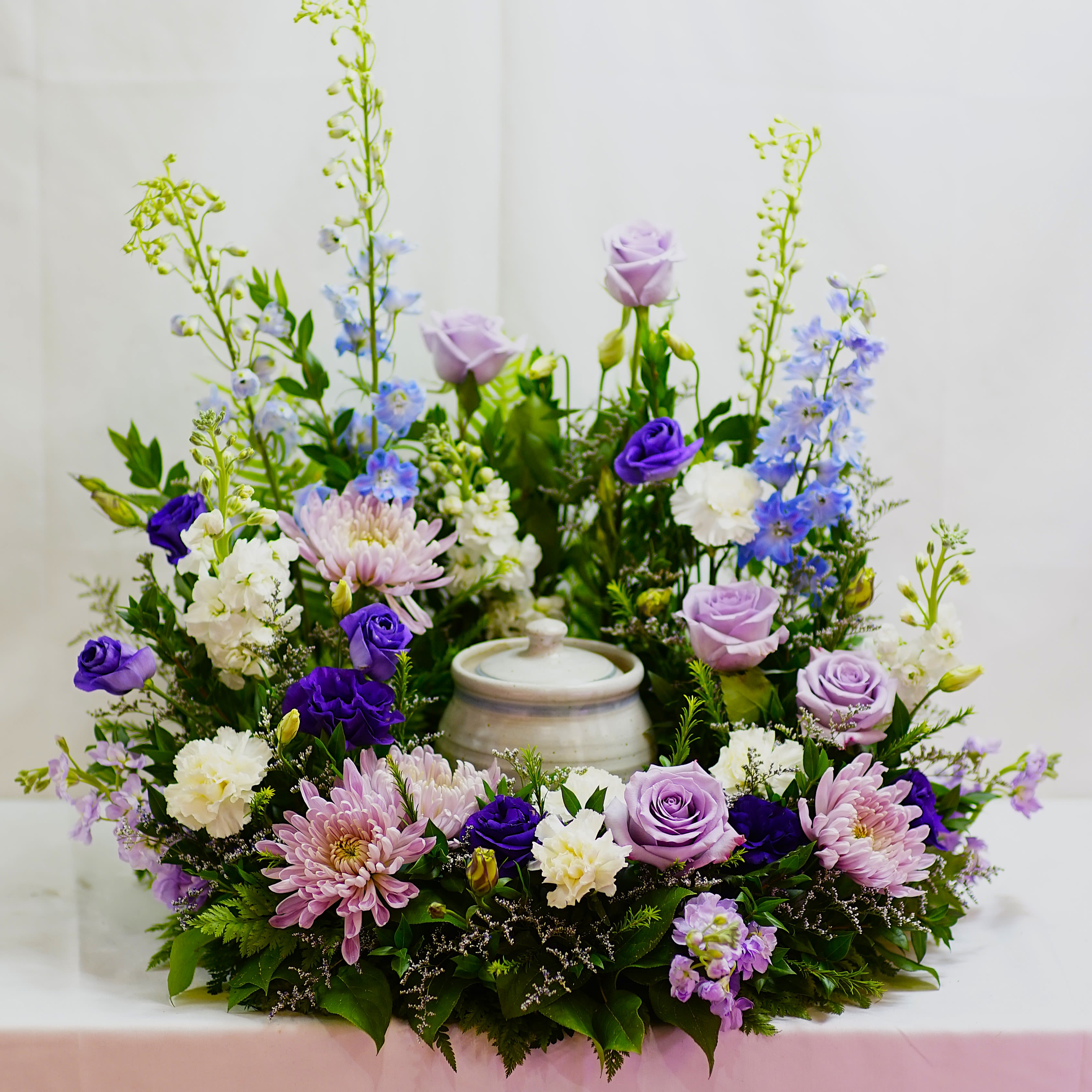 Twilight Garden Urn Tribute - A serene and elegant display, the Twilight Garden Urn Tribute is designed to surround the urn with beauty, peace, and reverence. Featuring soft lavender roses, delicate blue delphinium, regal purple lisianthus, creamy white carnations, and blush chrysanthemums, this arrangement blends cool and calming tones with lush greenery for a dignified, heartfelt tribute.  Perfect for memorial services or celebration of life ceremonies, this design brings gentle comfort and grace to any setting. Custom color palettes are available upon request to reflect your loved one’s personality or favorite blooms, ensuring a truly personal expression of remembrance.