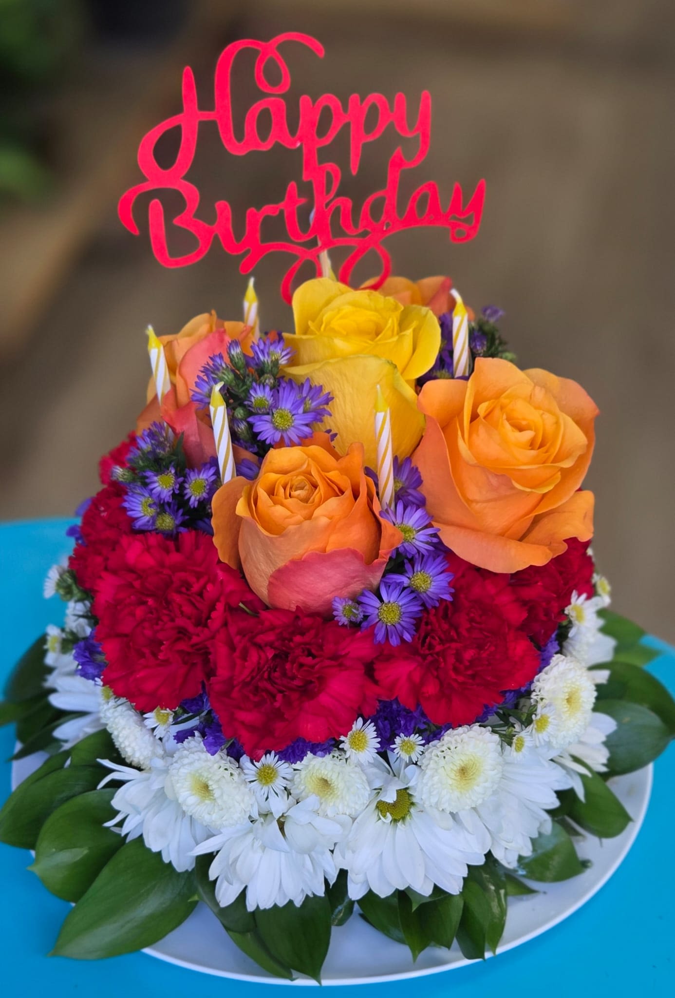 Happy Bloom Cake Happy Bloom Cake - A charming floral cake made with orange roses, red carnations, white e daisies , purple asters and lush greenery- beautifully arranged in a birthday cake shape. Incluide a"Happy Birthday " topper and decorative cnadles for a festuive and un forgettable touch. Perfect for celebrations , surprise gifts and original centerpiece arrangements.