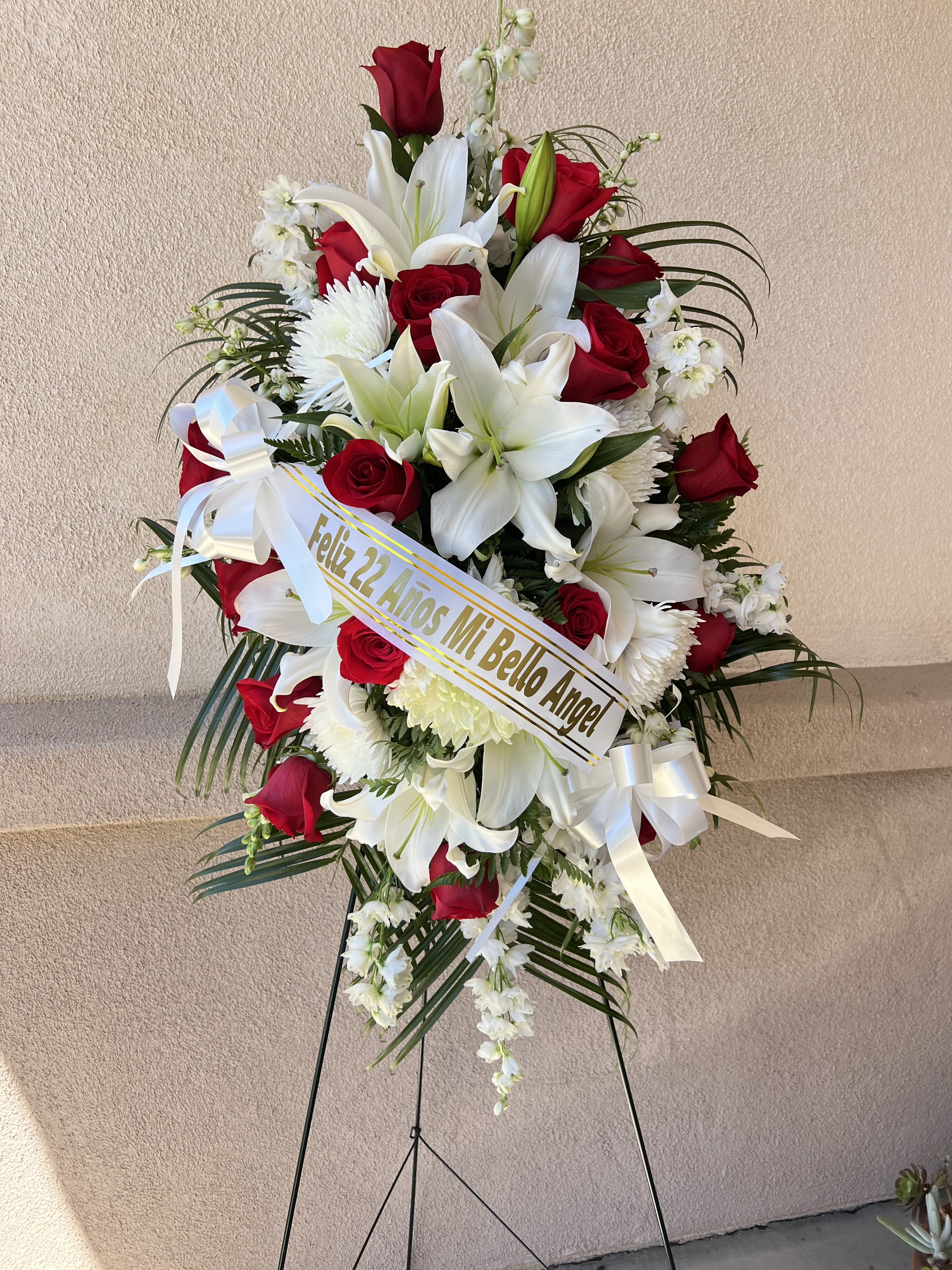 Eternal Grace Standing Spray  - A touching tribute of love and remembrance, the Eternal Grace Standing Spray is a graceful display of fresh blooms artfully arranged to offer comfort and honor a cherished life. Designed on an easel, it features a harmonious blend of flowers , white lilies, red roses, and lush greenery. symbolizing peace, respect, and eternal love. Perfect for funeral services, memorials, or graveside tributes. 