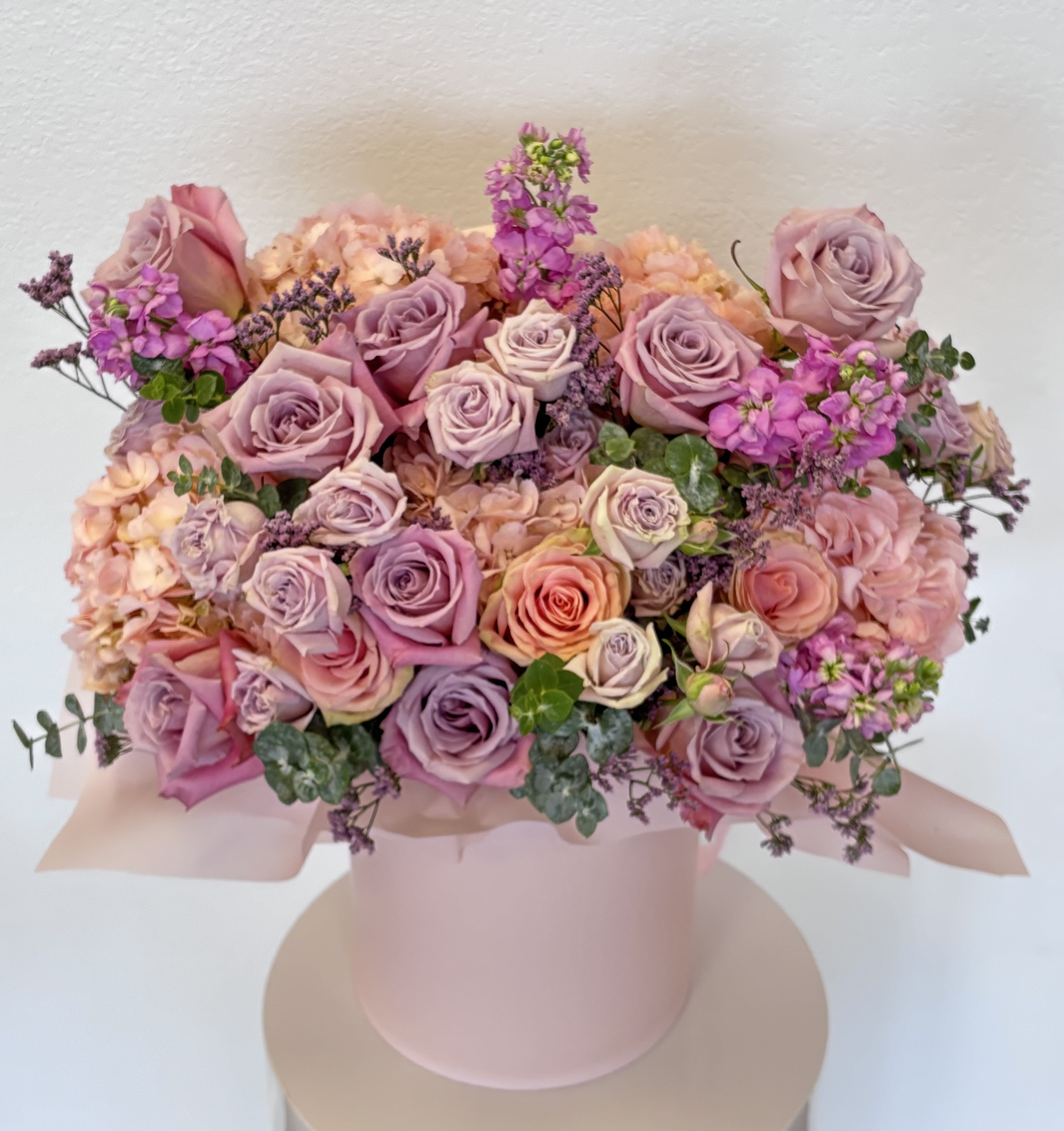 Château Bloom - An opulent floral statement inspired by the gardens of a French château. Château Bloom features a decadent blend of blush, lavender, and peach roses, accented with lush hydrangeas, mauve stock, and trailing eucalyptus. Arranged in a soft pink hatbox, this bouquet exudes timeless romance and refined elegance—perfect for milestone celebrations or unforgettable gifting moments.
