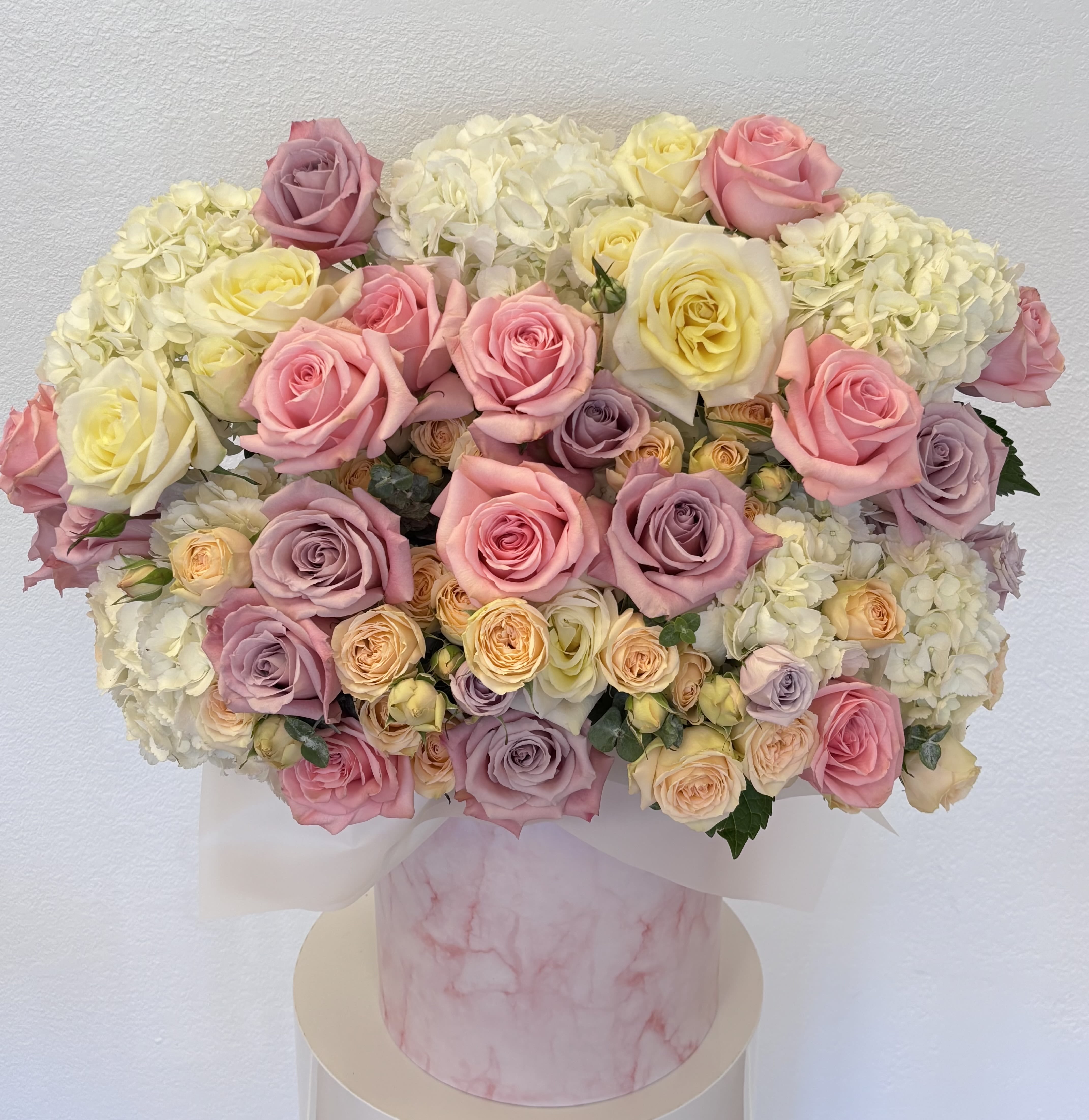 Versailles Dawn Versailles Dawn - A radiant masterpiece inspired by the early light over the gardens of Versailles. Versailles Dawn showcases a regal medley of ivory hydrangeas, buttery yellow roses, soft blush pinks, and lilac-toned blooms—all arranged in a luxurious pink marble hatbox. Elegant, airy, and breathtakingly romantic, this bouquet brings a sense of nobility and refined joy to any occasion.
