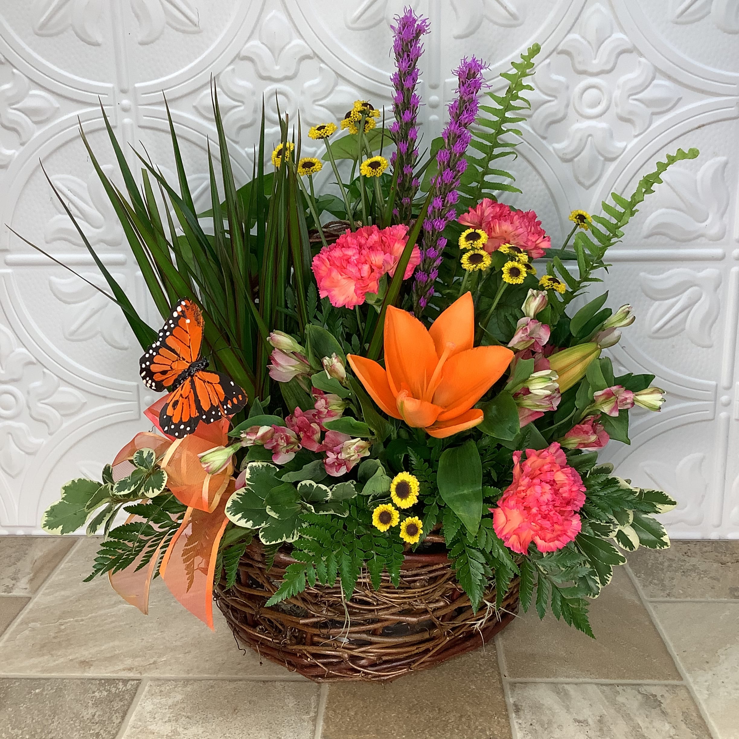 Marci’s Garden Marci’s Garden - Marci’s Garden is a lovely combination of live plants and fresh cut flowers in a wicker basket. It includes two green houseplants along with an arrangement of orange asiatic lilies, carnations, charmelia, liatris, yellow button poms and other seasonal blooms and additional greens. A butterfly floral pick adds a special touch. Approximate size: 17” wide x 20” tall. Primary flowers: plant, carnation, lily, mum