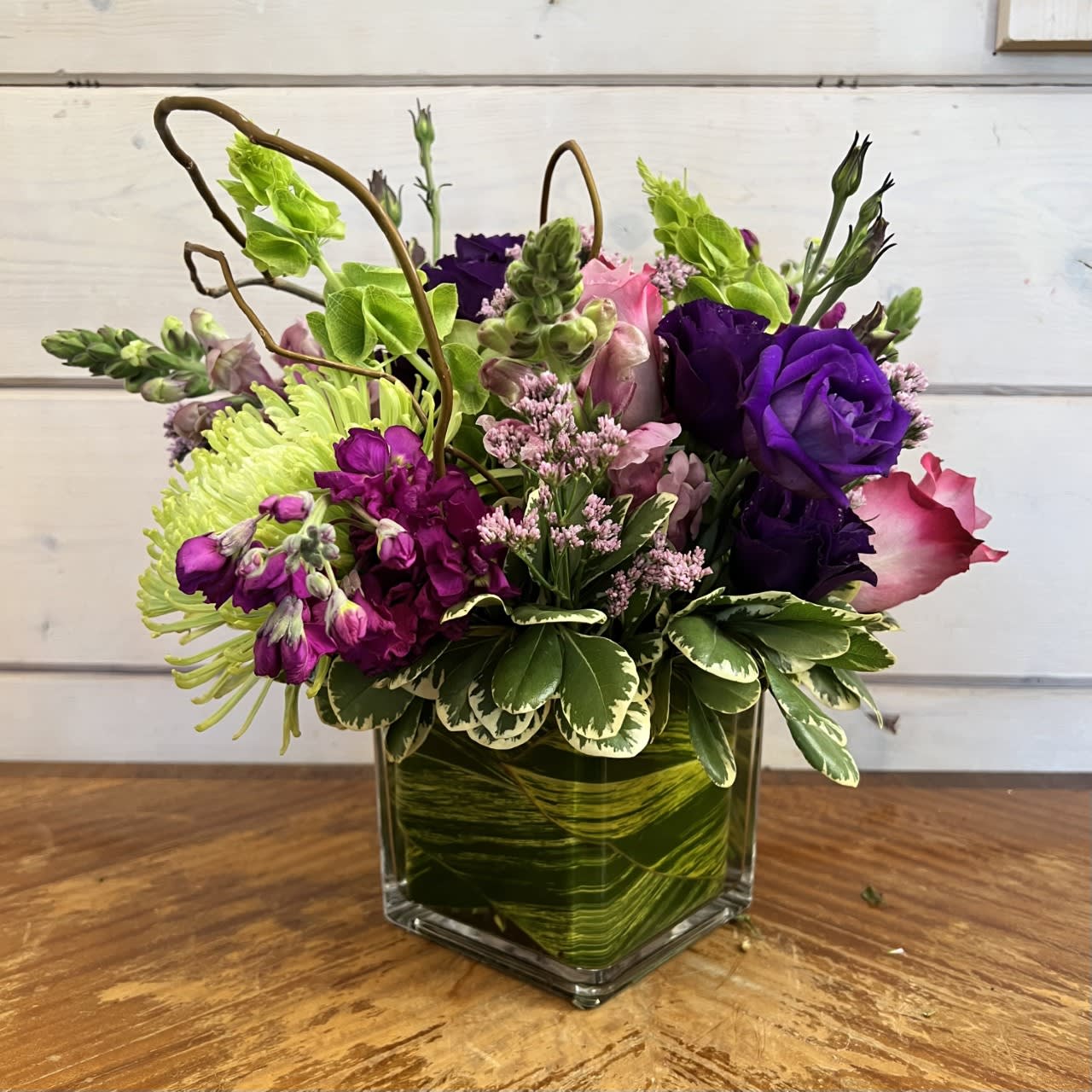 The Purple Paradise Bouquet The Purple Paradise Bouquet - Lavender roses, purple stock, purple lisianthus, lavender snapdragons, green spider chrysanthemums, bells of Ireland arranged in a modern 5in leaf lined cube with looped curly willow and lavender misty.