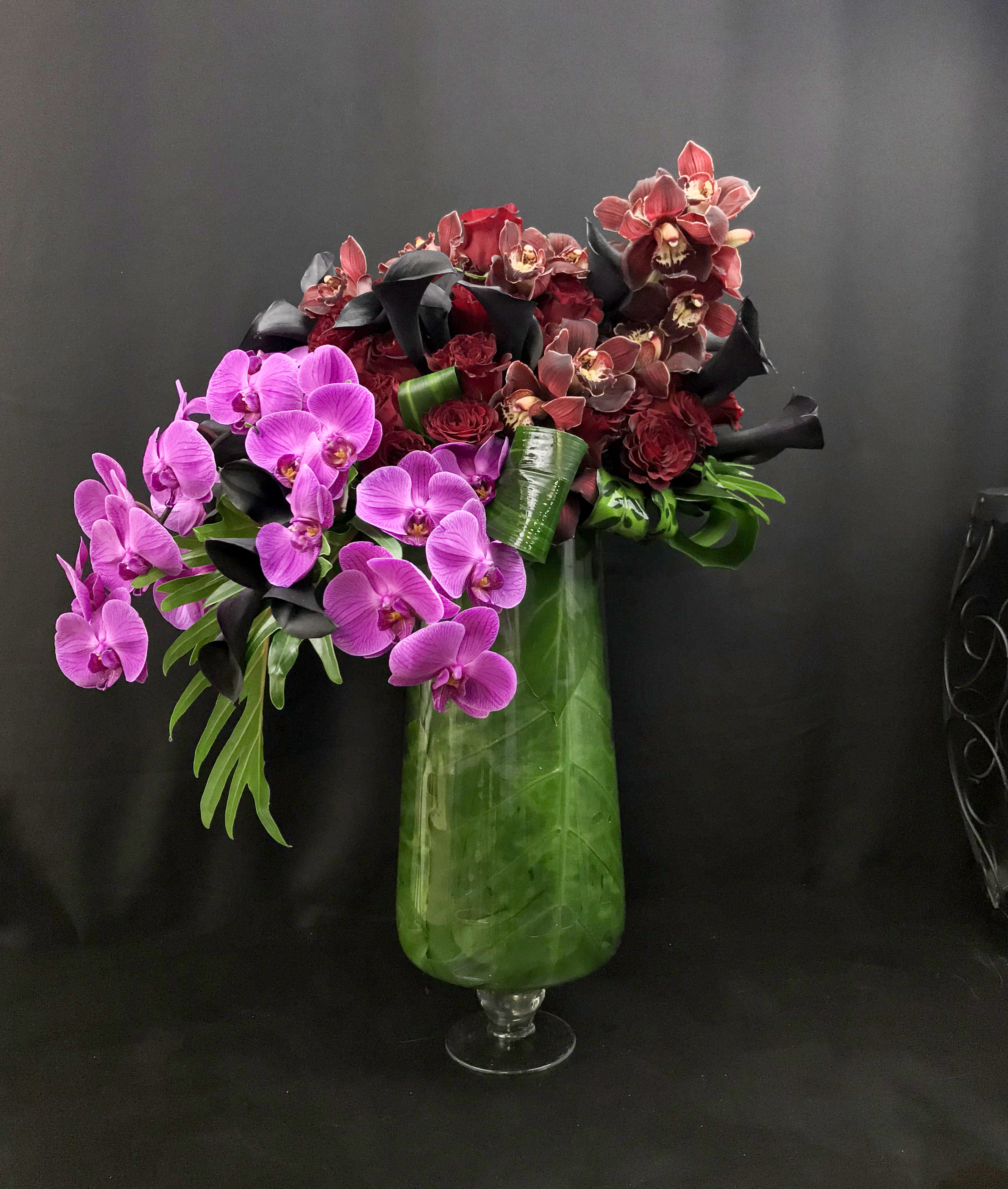 Orchids fantasia  - Tall, modern design with Phalaenopsis and cymbidium orchids, dark purple ( black) calla lilies and roses.  Arranged into clear vase with leaves decor