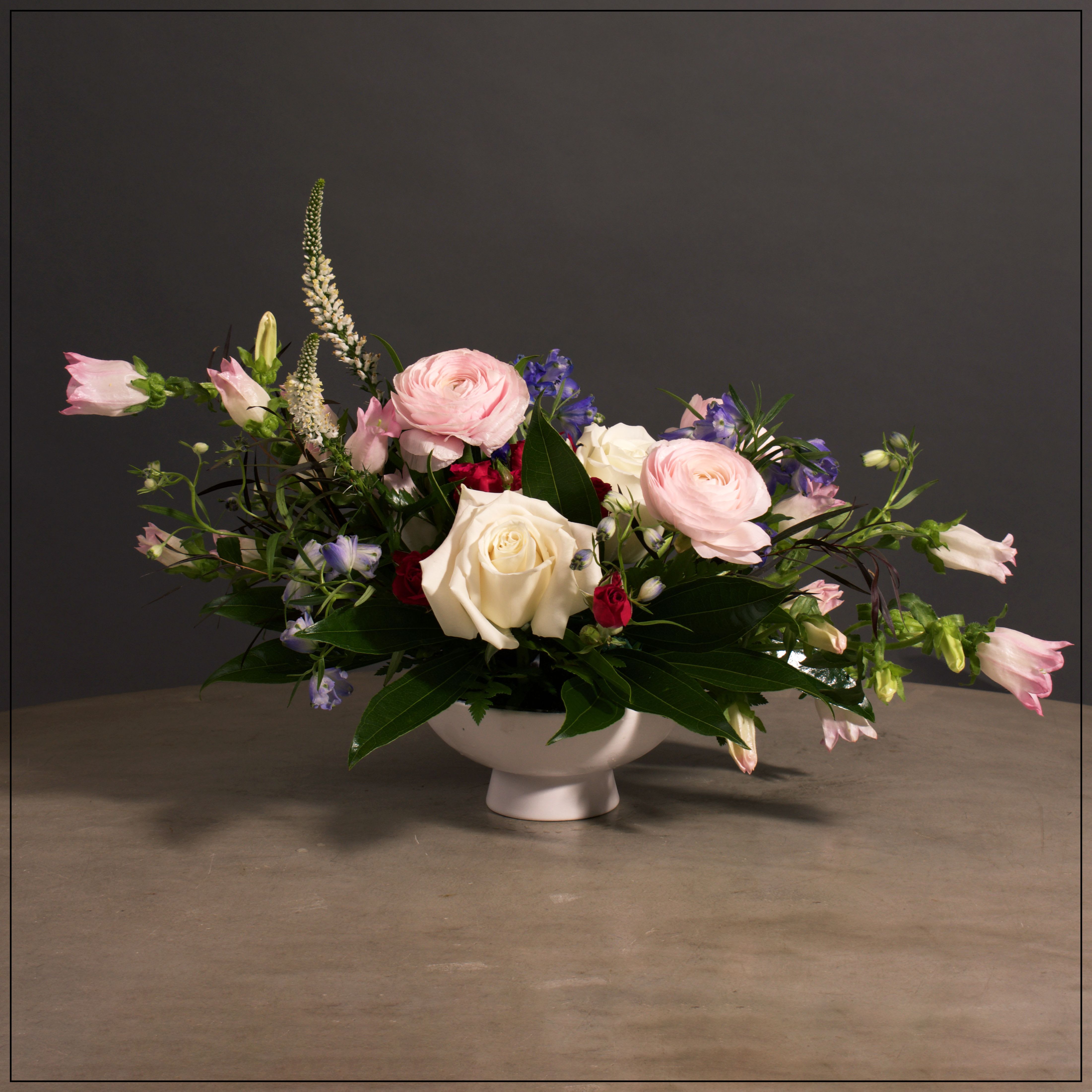 Eloise - Gorgeous modern design in a white Demi Bowl, featuring roses, spray roses, ranunculus, campanula, veronica, and delphinium.