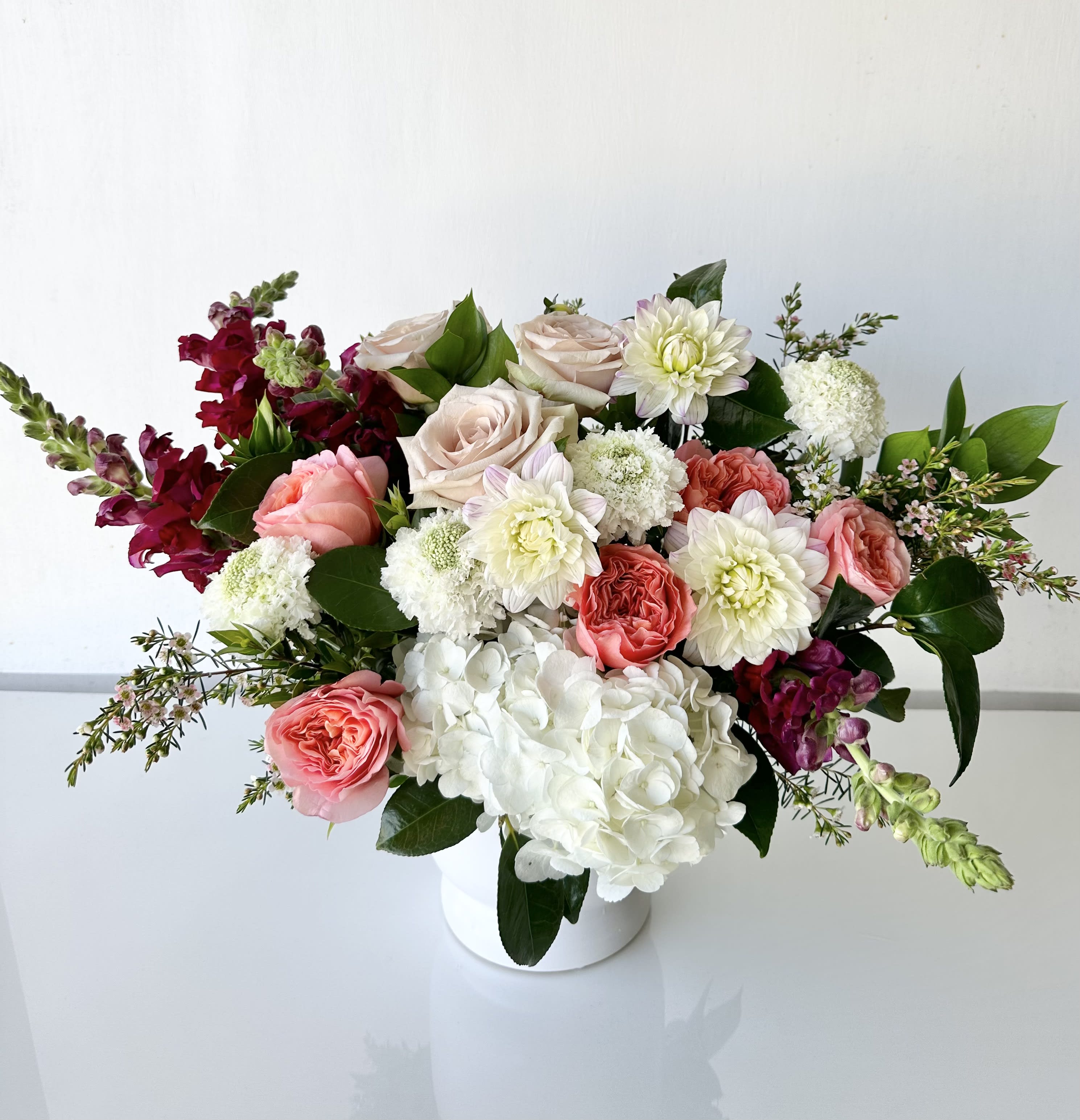 Symphony of Love - EFS - An arrangement of fresh-cut flowers includes roses, dahlia, scabiosa, hydrangea, and snapdragon in a white ceramic vase. Dimensions: 17&quot; H x 23&quot; W