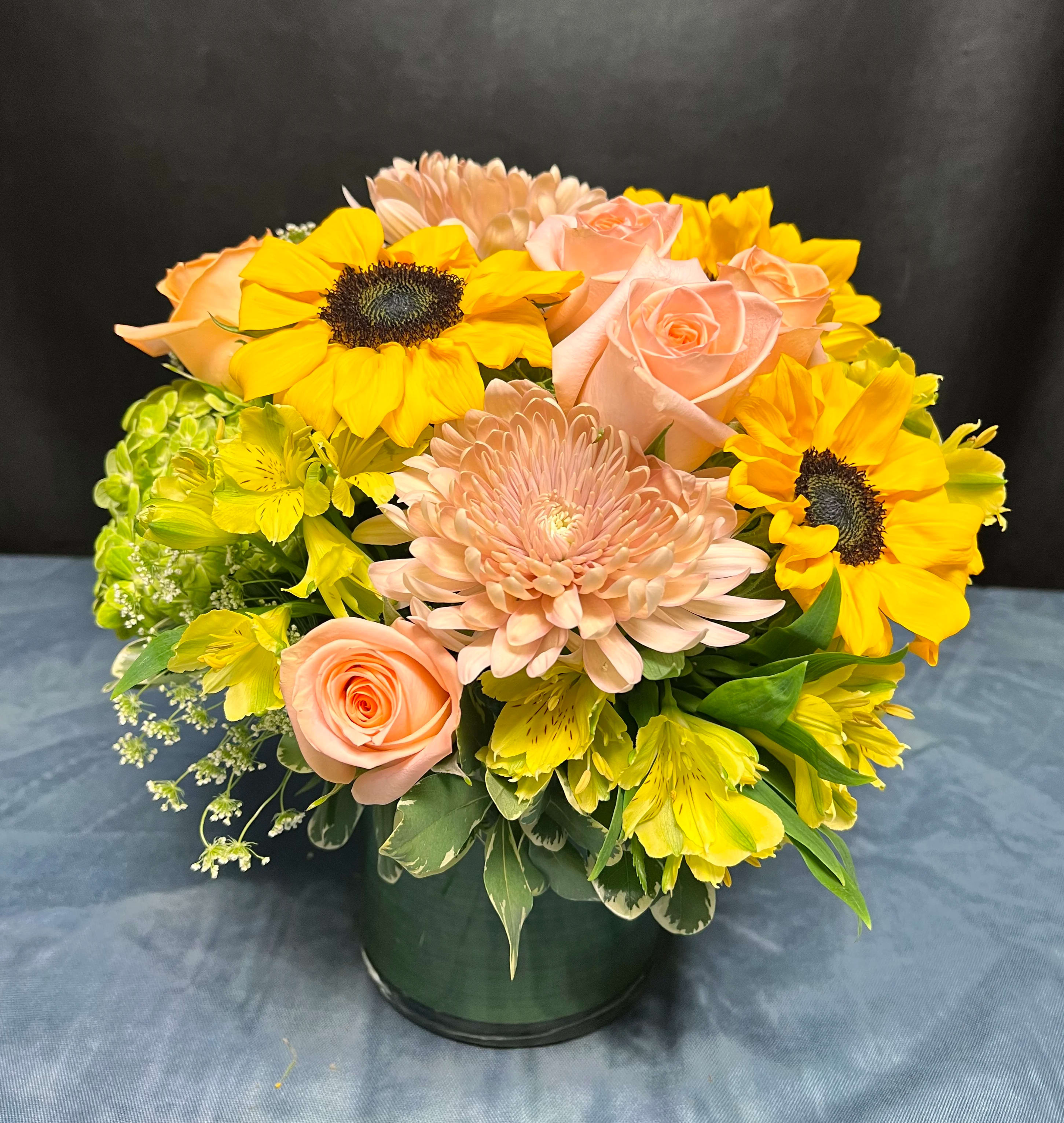 First Light  - Like the gentle glow of morning, this arrangement radiates warmth and optimism. Featuring soft peach roses, golden sunflowers, delicate queen Anne’s lace, vibrant alstroemerias, and charming peach mums, all accented with fresh greenery. First Light is a celebration of new beginnings, designed to brighten any space with natural beauty and heartfelt cheer.