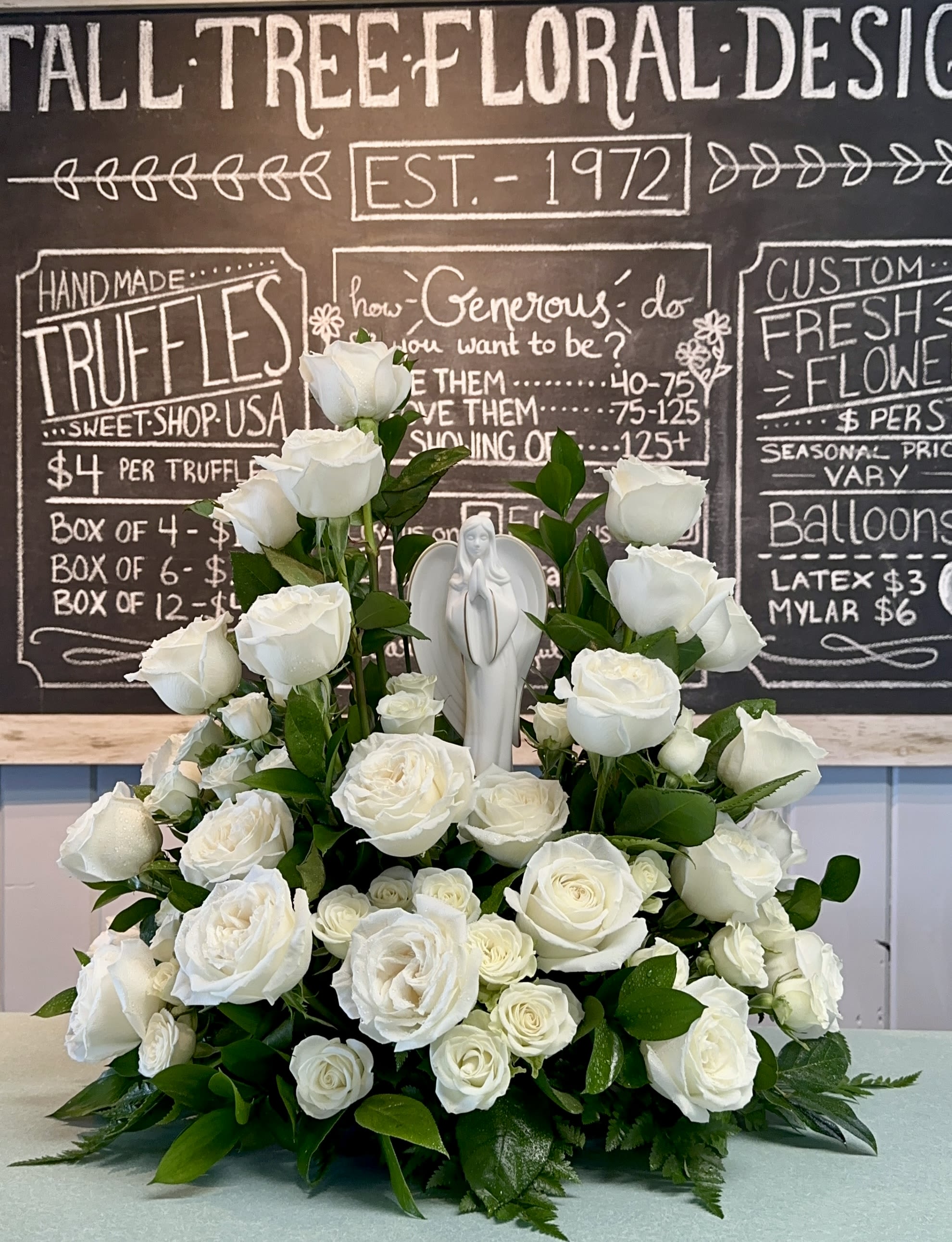 Heavenly Grace - A serene tribute of pure white roses and lush greenery, crowned by a graceful Madonna Statue.