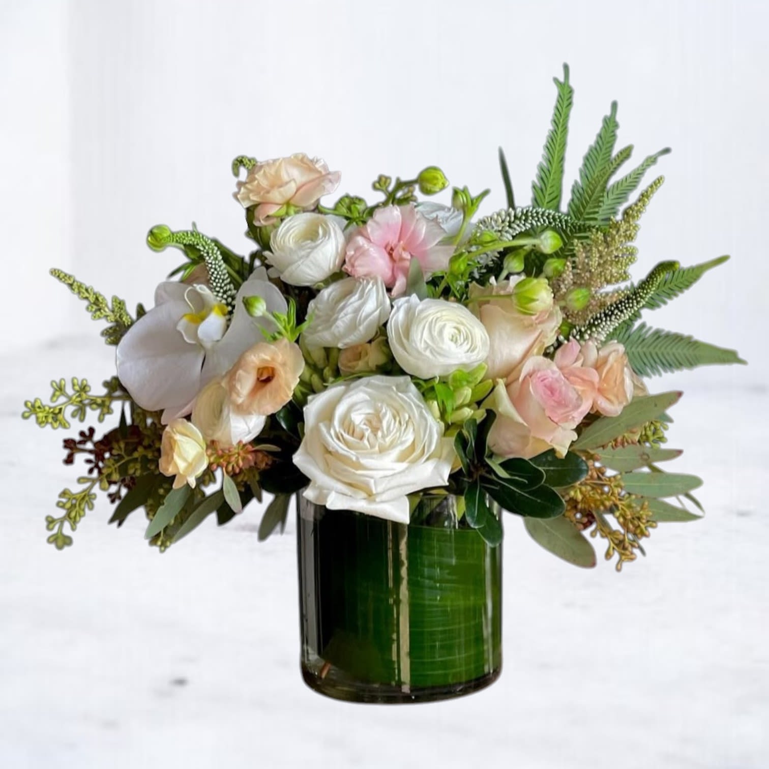 Silken Still - Understated and serene, Silken Still is a refined floral arrangement that whispers elegance. This peaceful design features creamy white roses, pale blush lisianthus, ranunculus, orchids, and delicate greenery—gracefully arranged in a leaf-wrapped glass vessel. The soft textures and gentle tones create a sense of quiet luxury and effortless calm.