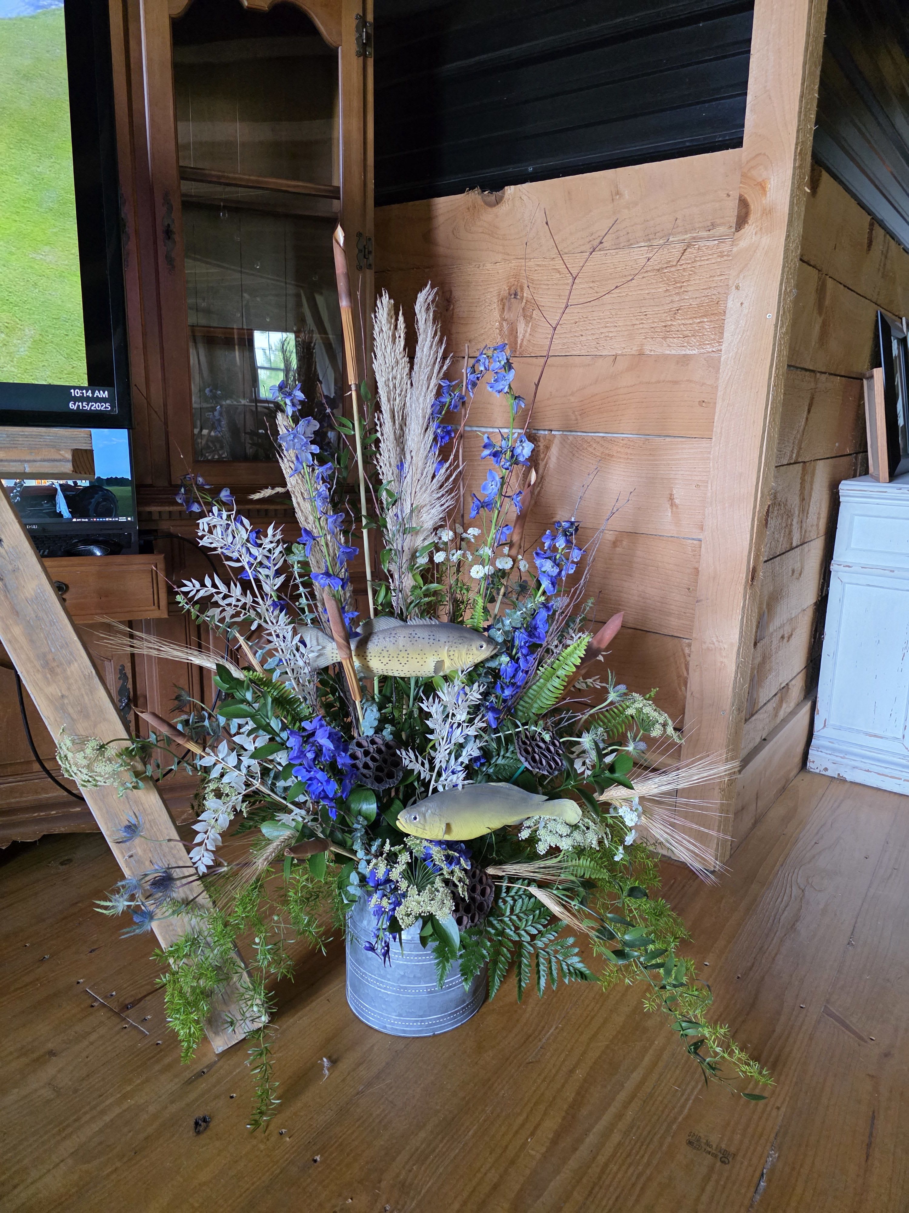 The Fisherman - For the man who loved to fish, this stunning display of blue delphinium, mixed seasonal greenery, dried grasses and dried foliage, create a dramatic background for our beautiful simulated fish. Standing approximately 42&quot; tall and 3&quot; wide The Fisherman makes an impression.