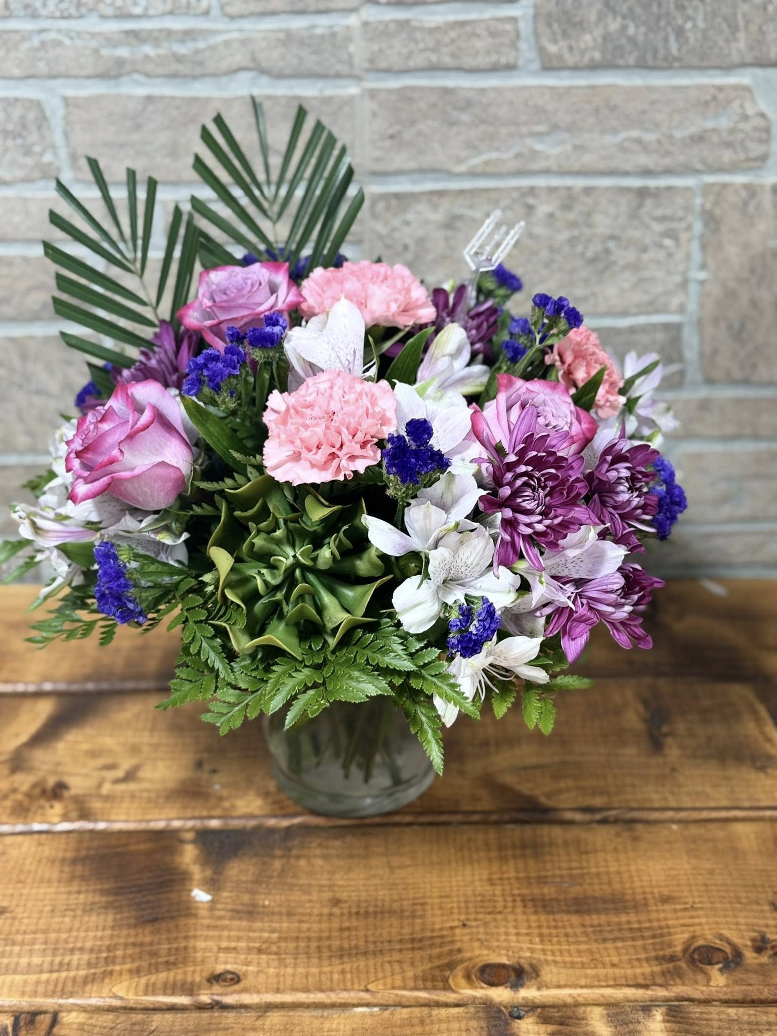 Whispers of Grace Whispers of Grace - Delicate, dreamy, and full of quiet beauty, Whispers of Grace is a soft symphony of purples and pinks. Featuring enchanting purple roses, blush pink carnations, lavender mums, purple alstroemeria, and airy purple statice, this arrangement is accented with lush robellini fern and a charming succulent for a modern touch. Thoughtfully designed to offer comfort, admiration, or gentle encouragement, this bouquet speaks volumes without saying a word.